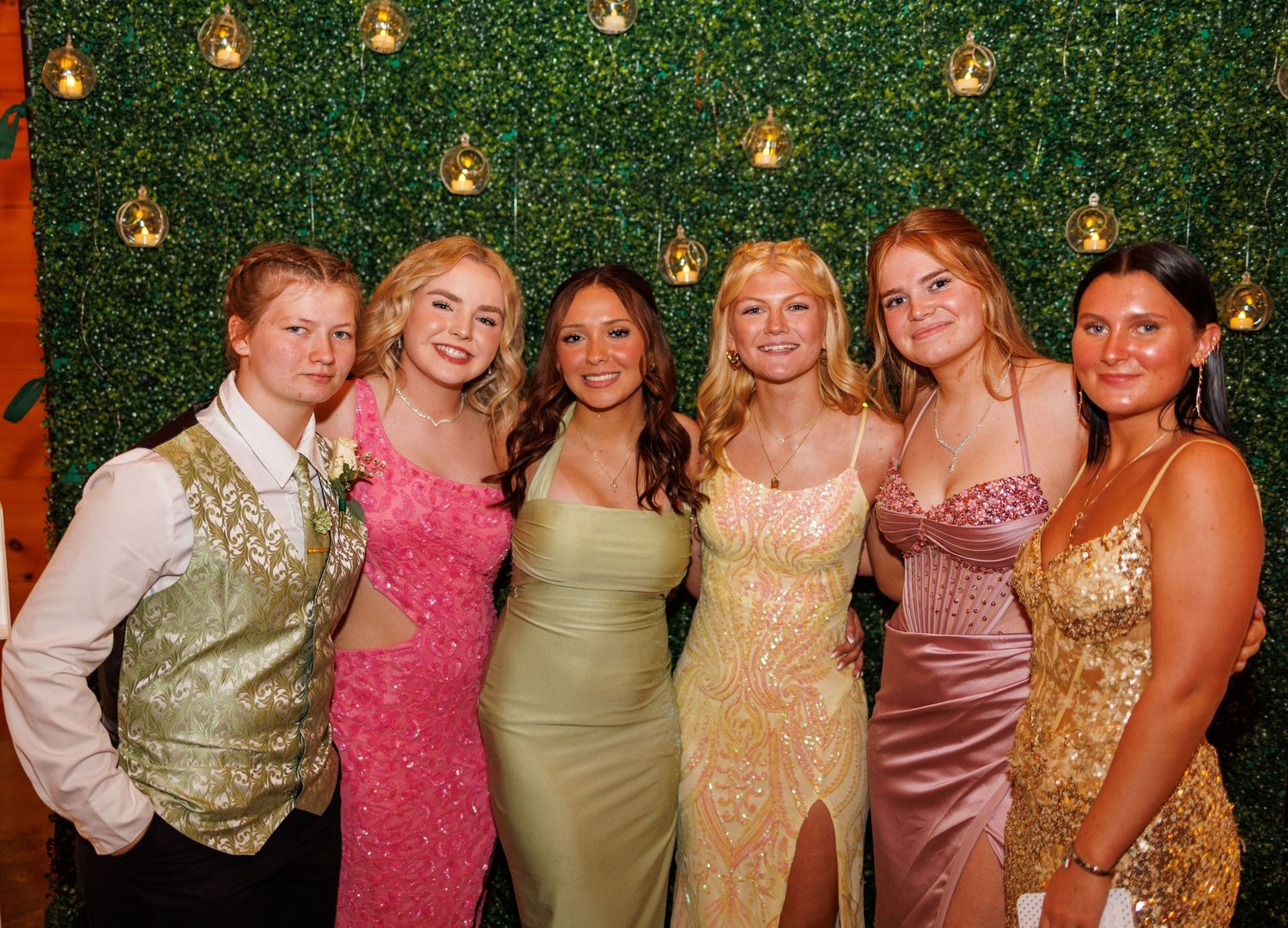 Six smiling students in formal prom attire posing together in front of a green hedge wall backdrop.