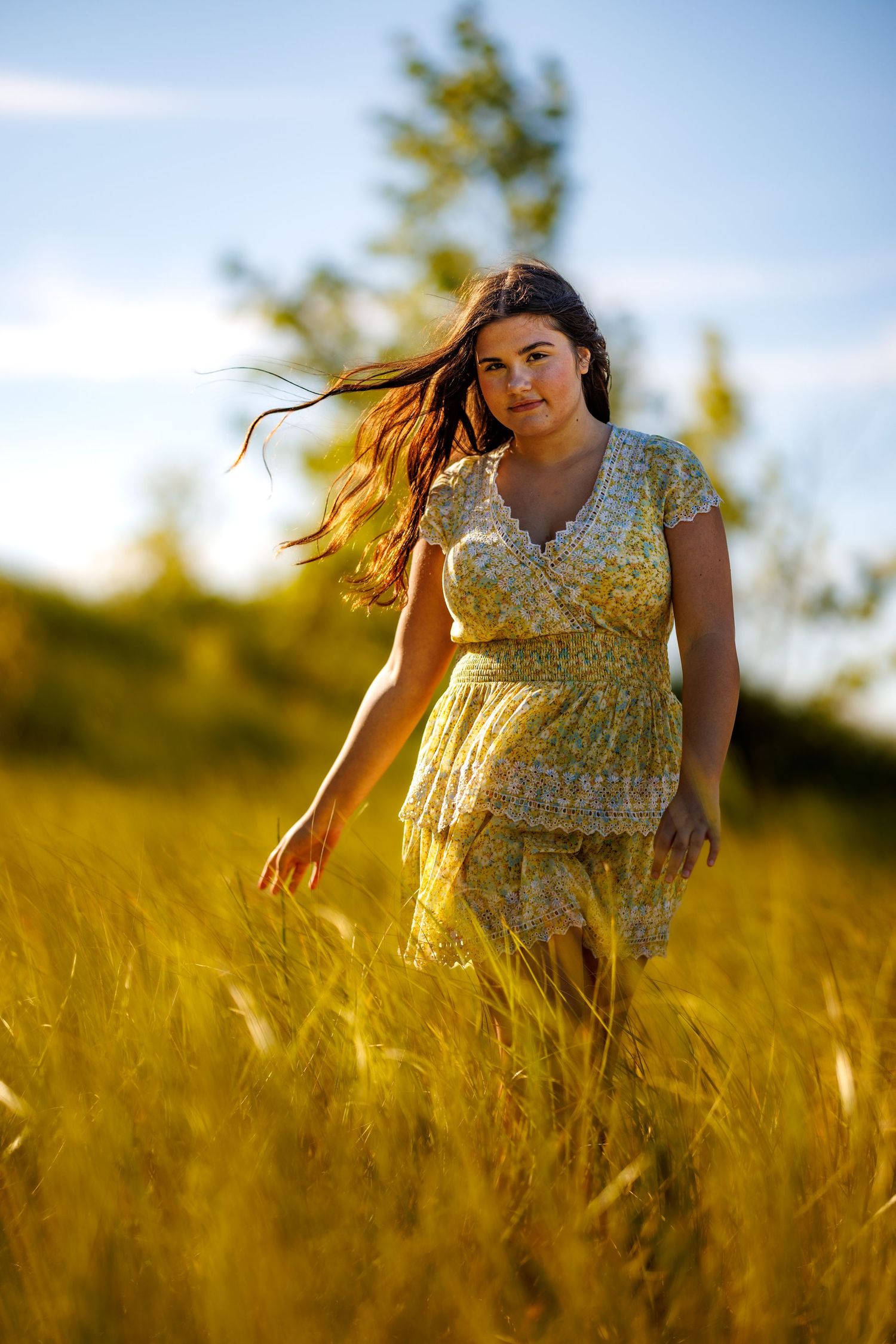 Young woman in yellow floral dress walking through golden wheat field at sunset with hair blowing in wind.
