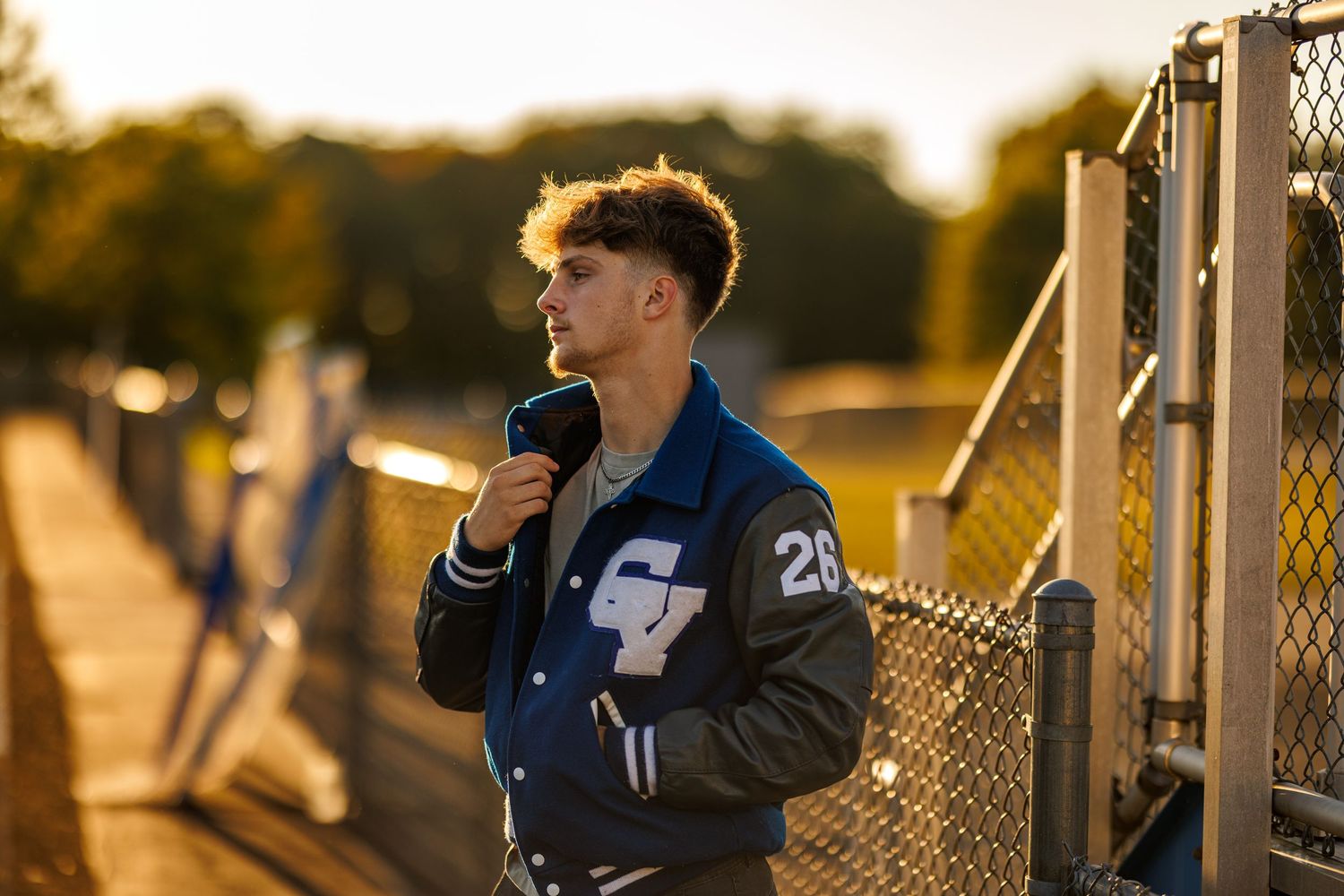 Young man in varsity jacket posing by a chain-link fence in golden light.