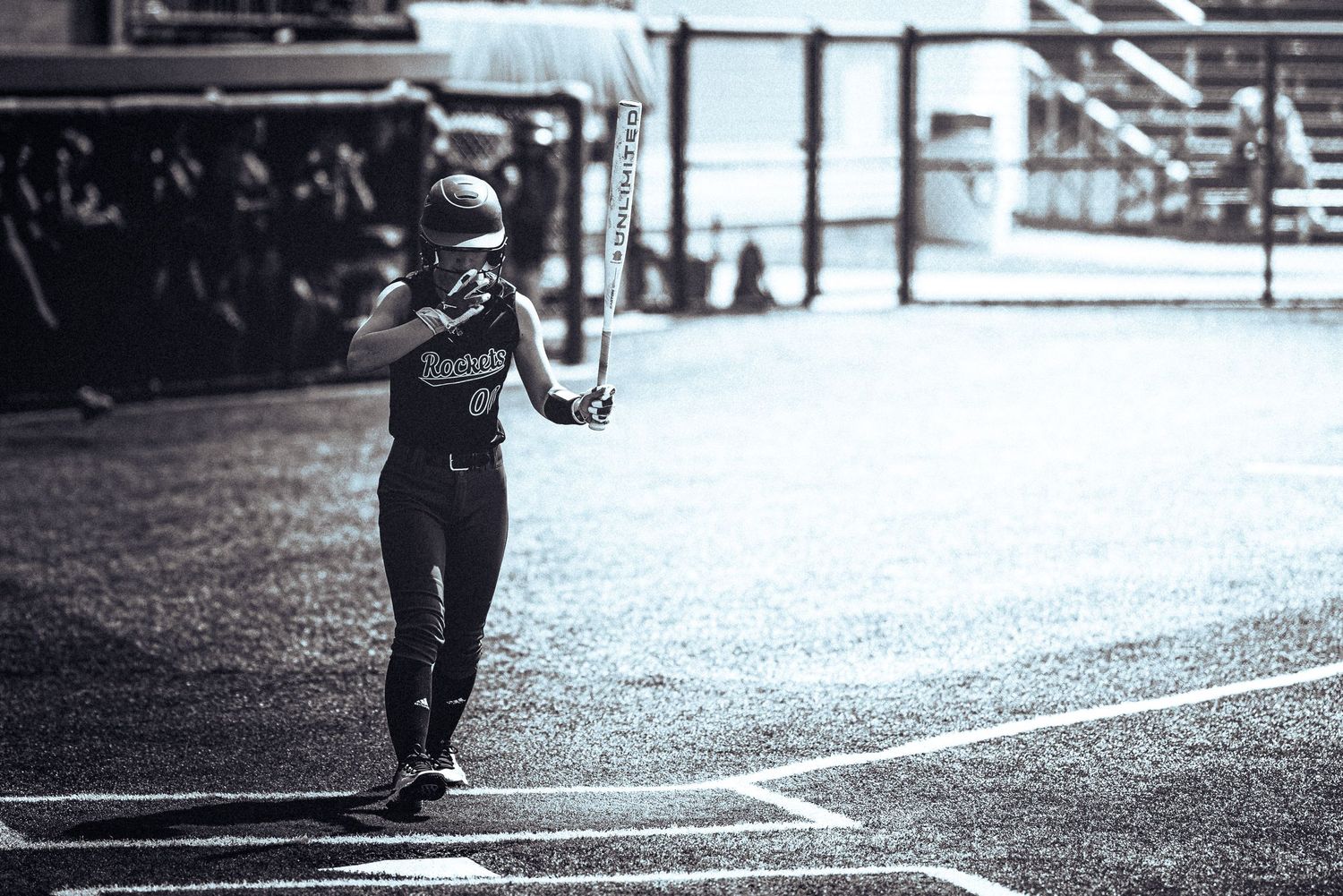 A softball player walks along the baseline carrying a bat in a dramatic black and white game photograph.