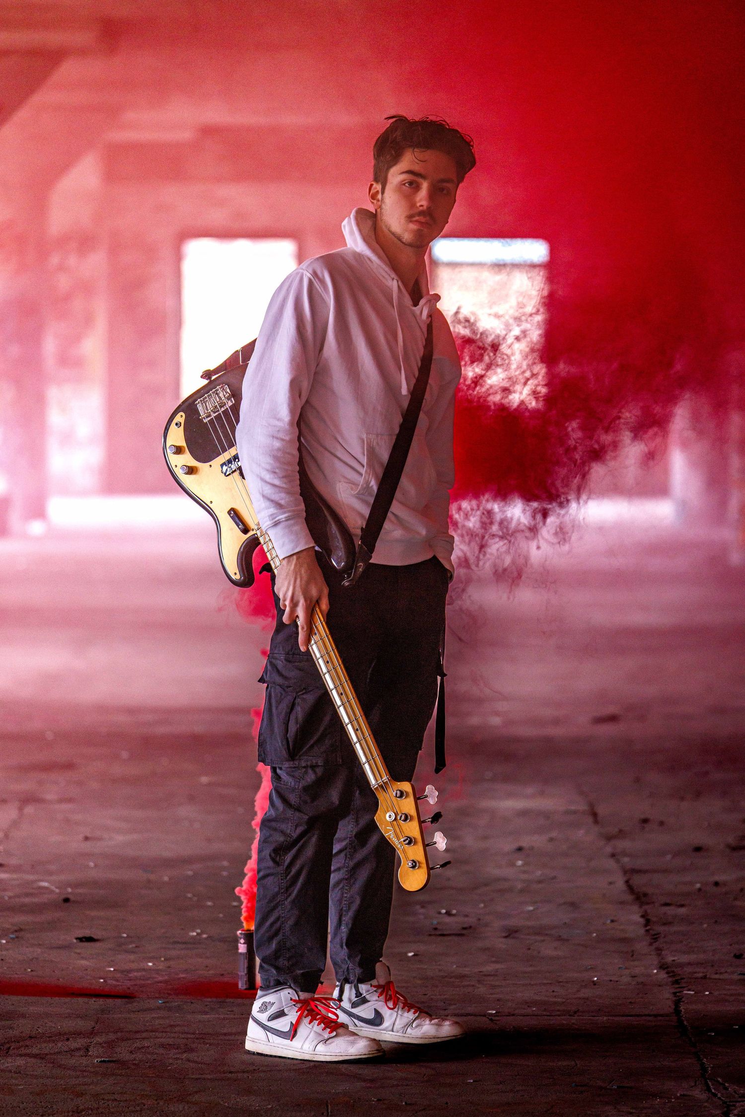 Guitarist in white shirt holds bass guitar in smoky red-lit abandoned space.