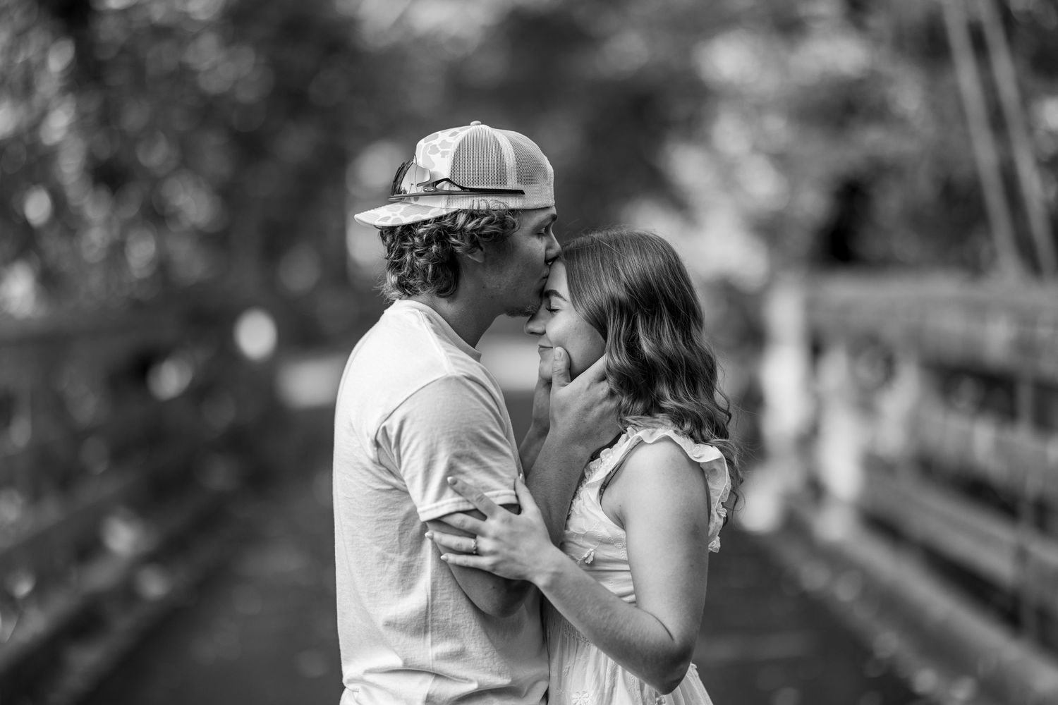 A black and white romantic photo of a young couple about to kiss on a wooden bridge outdoors.