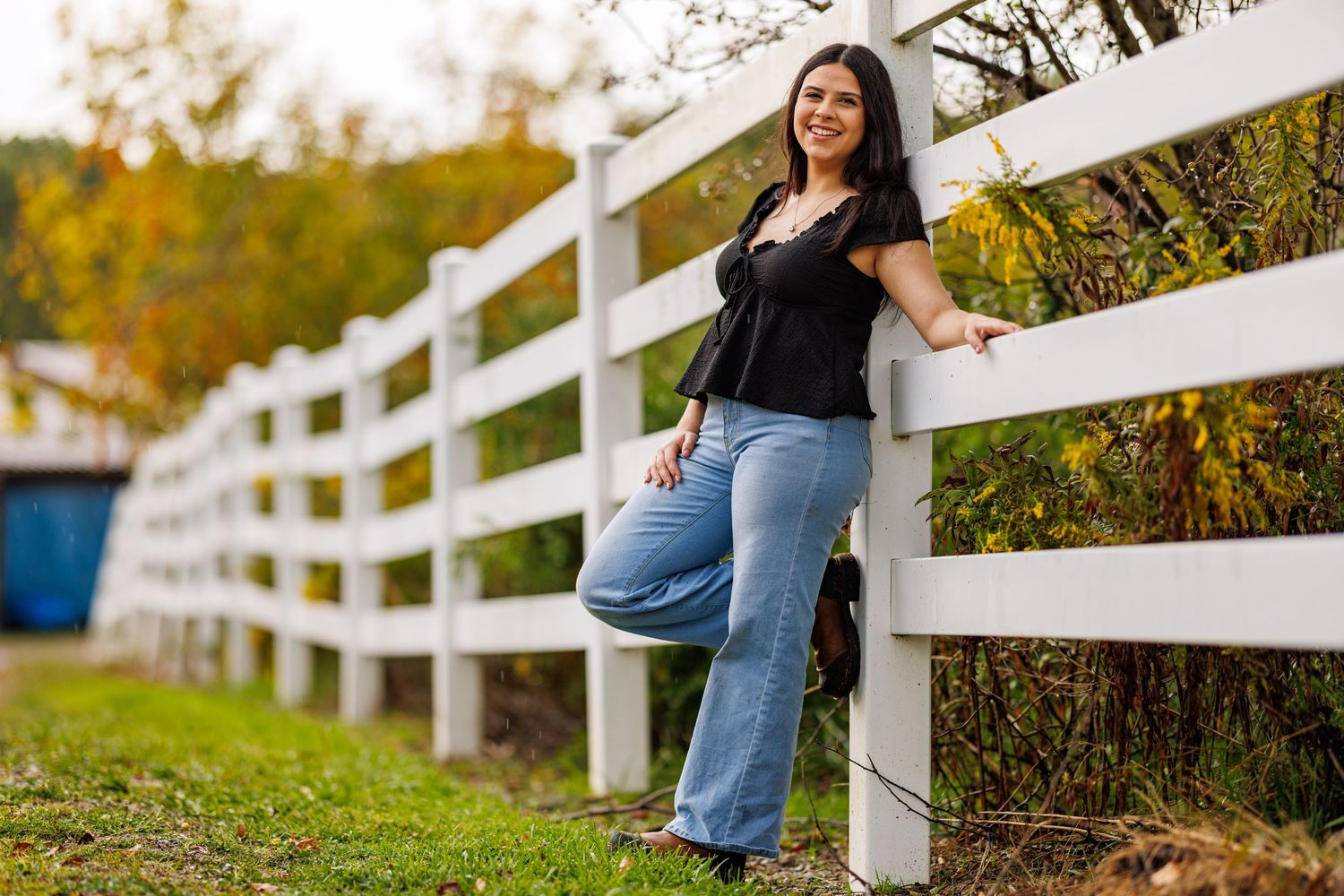 Young woman in black top and flared jeans leaning on white wooden fence outdoors.