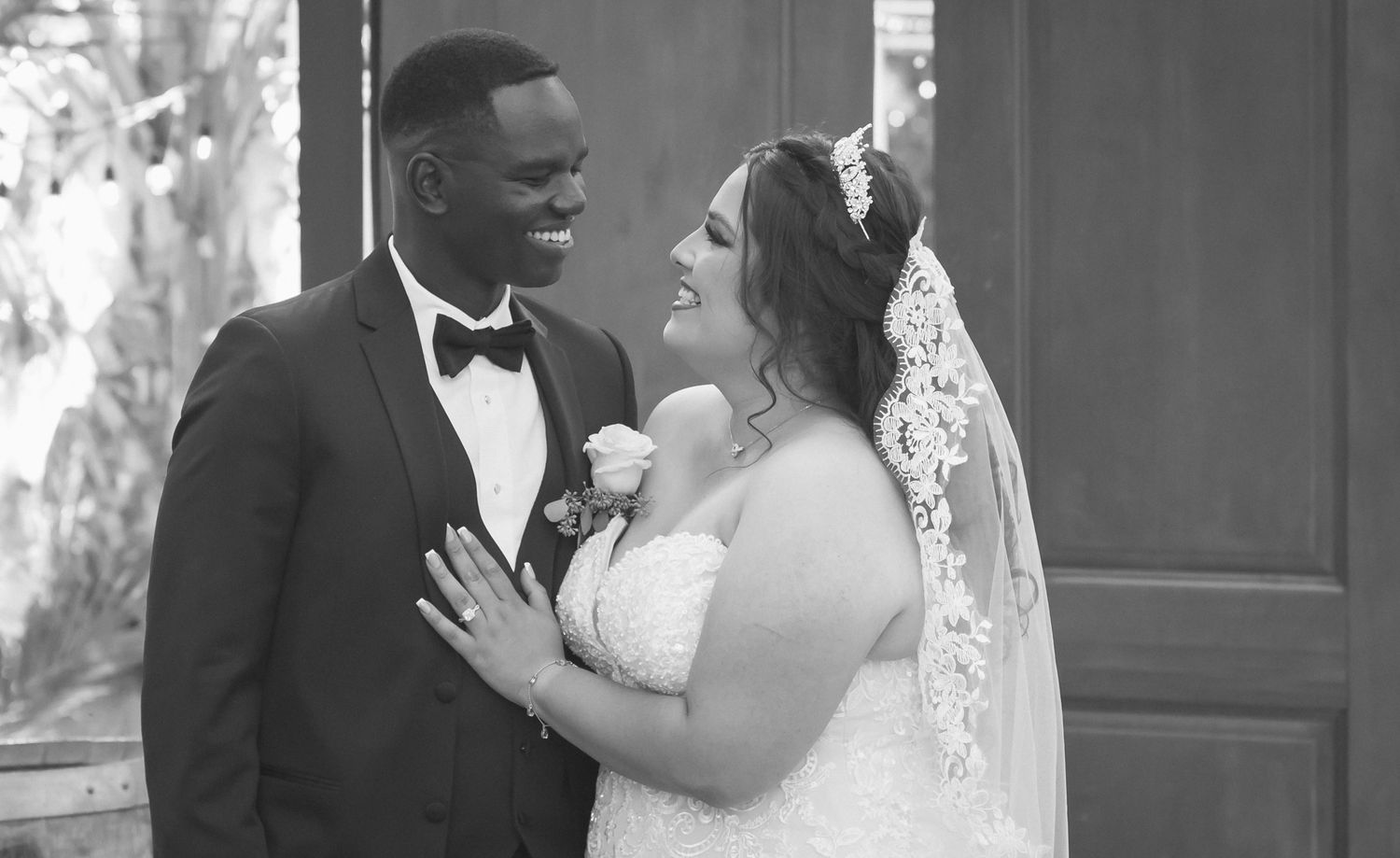 Black and white photo of a smiling bride and groom in wedding attire gazing at each other outdoors.