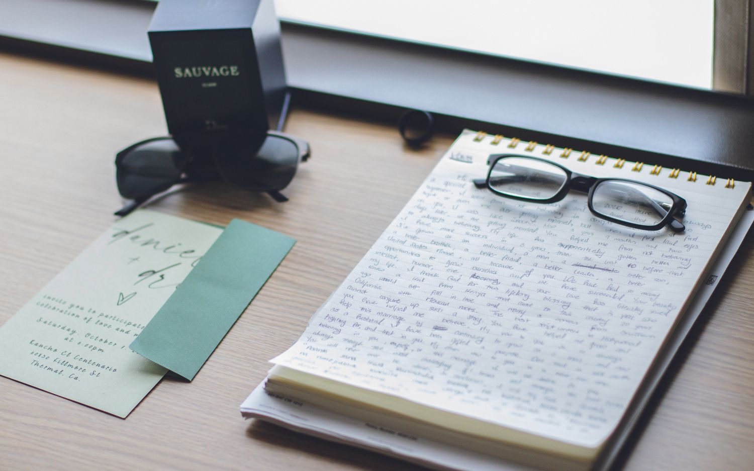 A desk with a spiral notebook, glasses, green card, sunglasses, and Sauvage cologne box on a wooden surface.