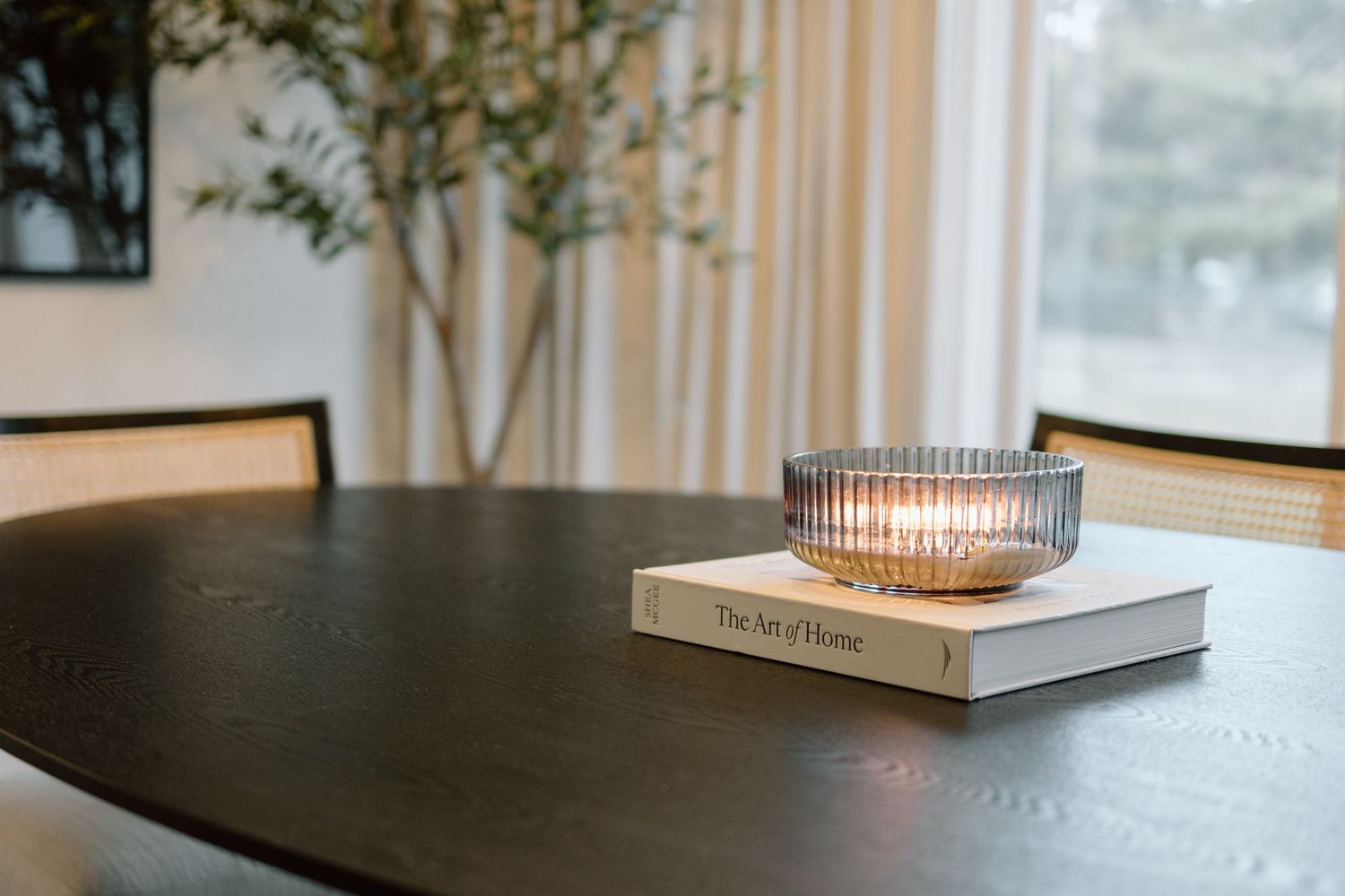 Dark wood round dining table with a book and lit glass candle holder centerpiece near a bright window.