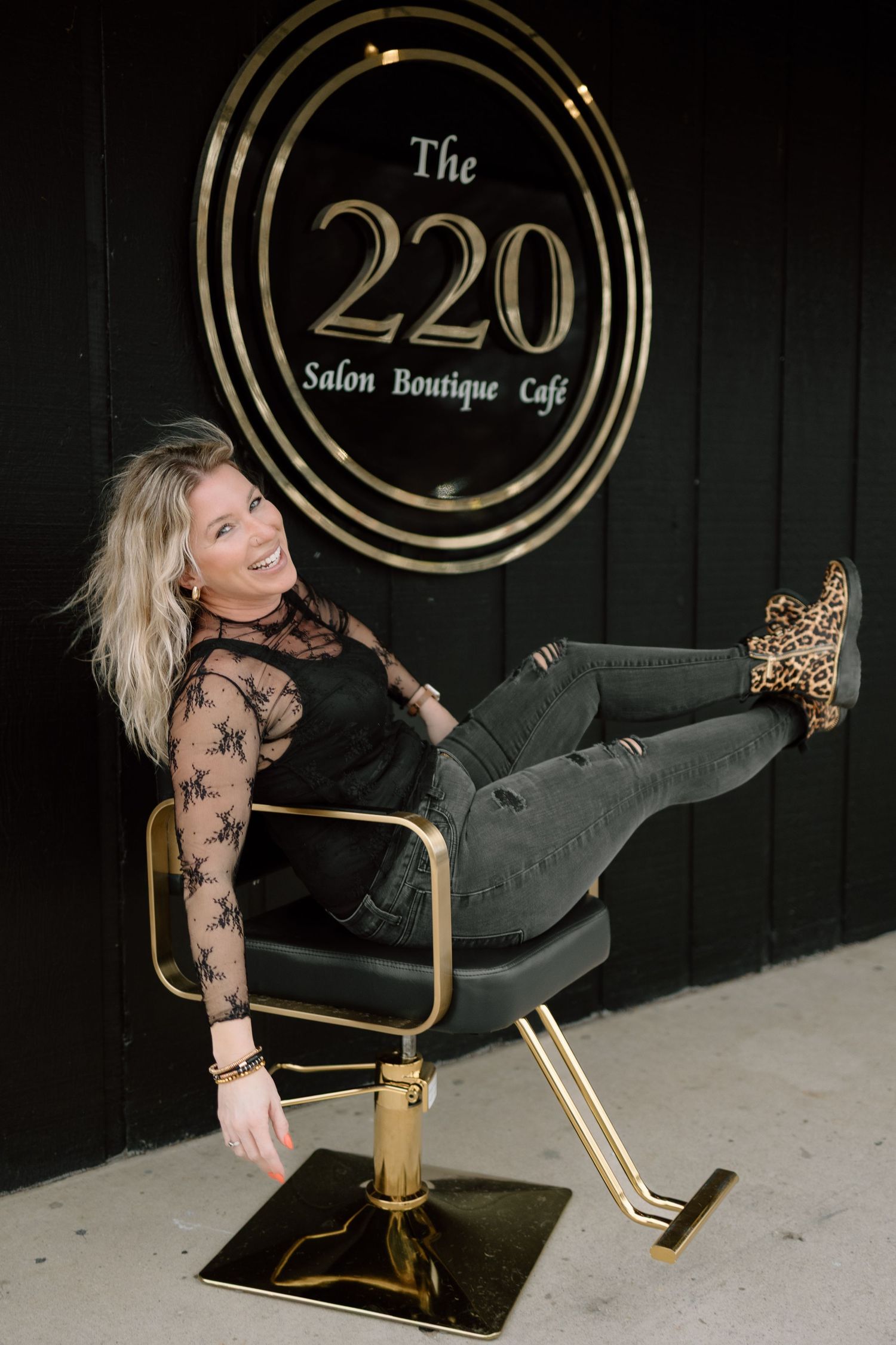 A smiling blonde woman sits in a gold salon chair in front of The 220 Salon Boutique sign.
