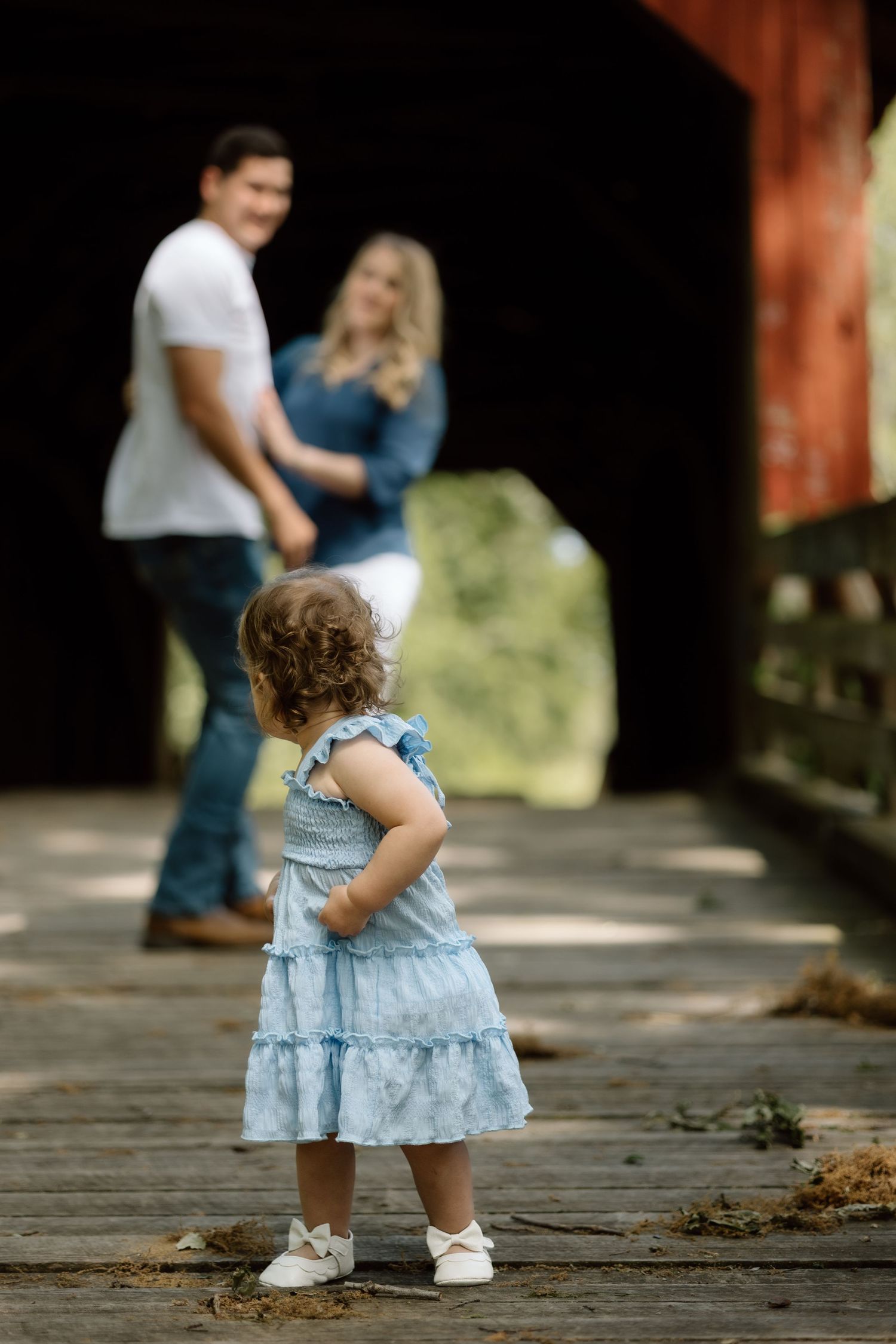 Toddler girl in blue dress watches her parents dance on a covered wooden bridge during a family photo session.