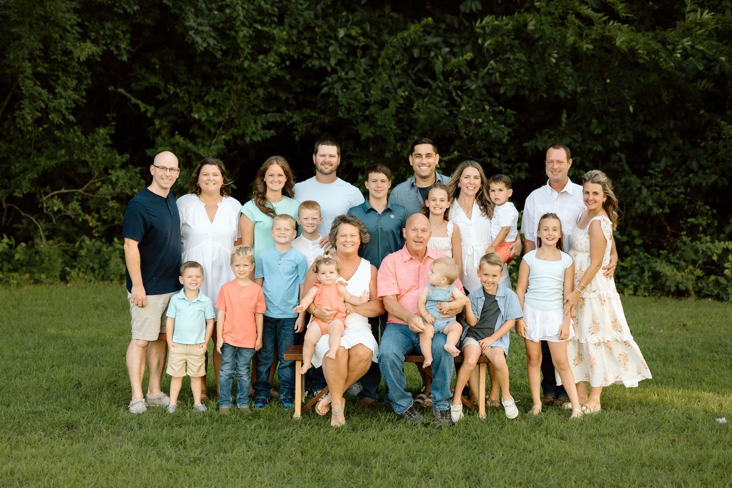 Large multigenerational family group portrait outdoors on green grass surrounded by lush summer trees.