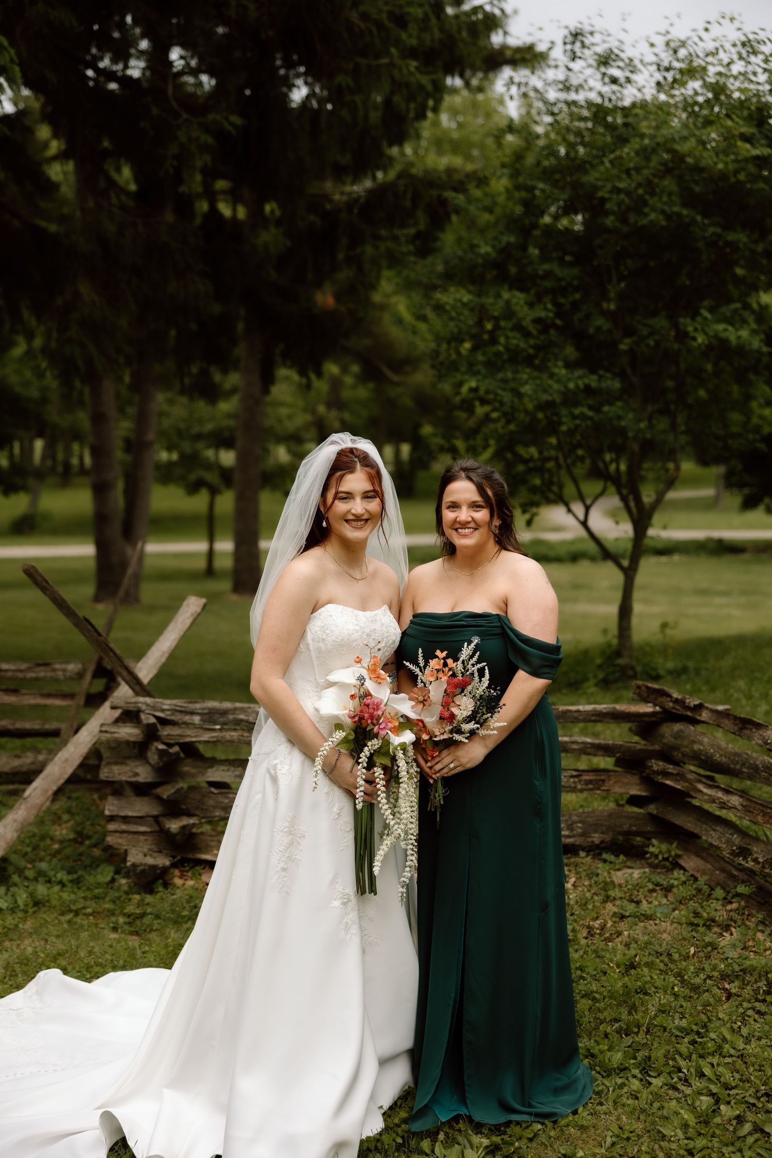 Bride in white strapless gown poses with bridesmaid in emerald off-shoulder dress outdoors.