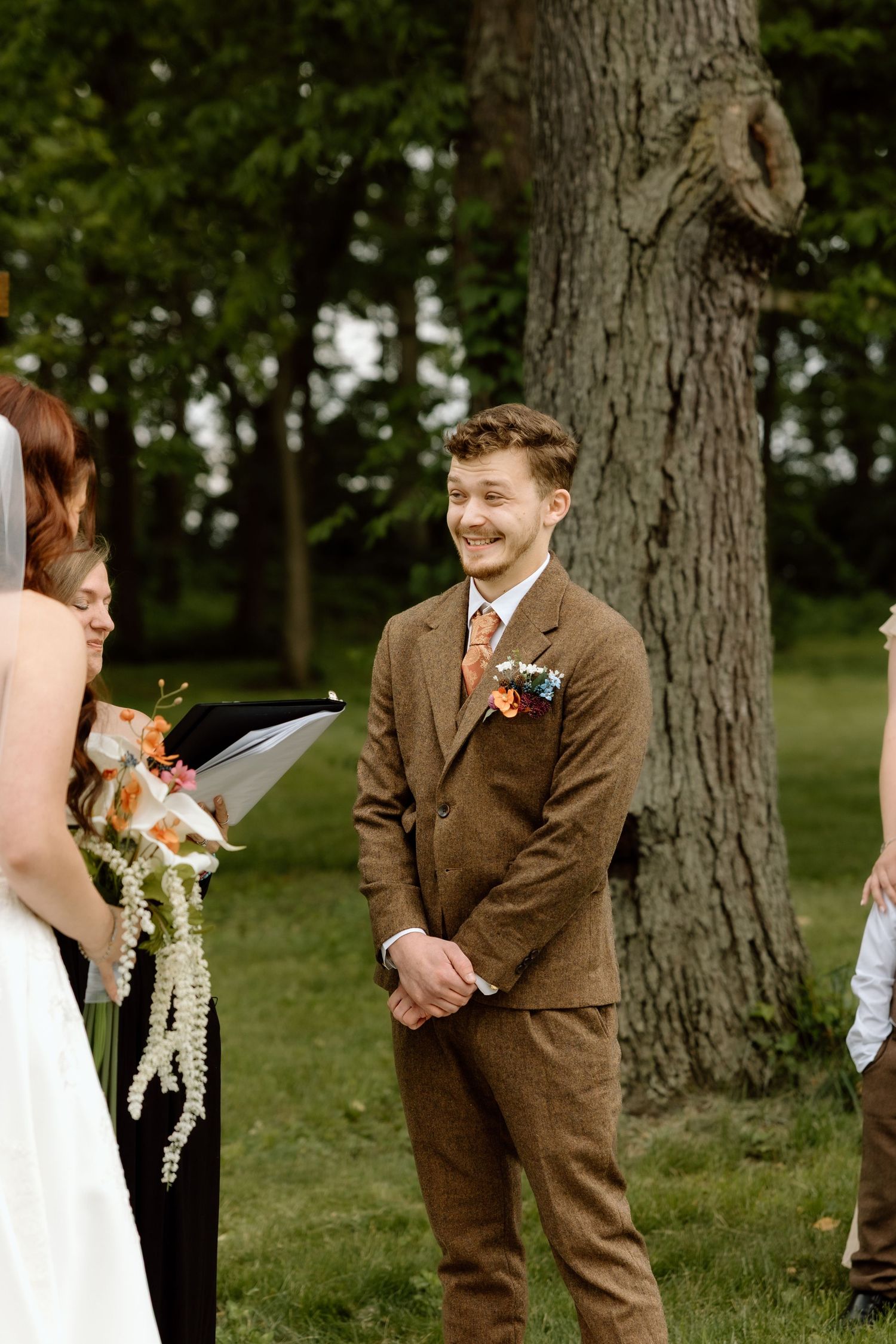 Groom in brown tweed suit smiles warmly at bride during outdoor wedding ceremony beneath tall oak trees.