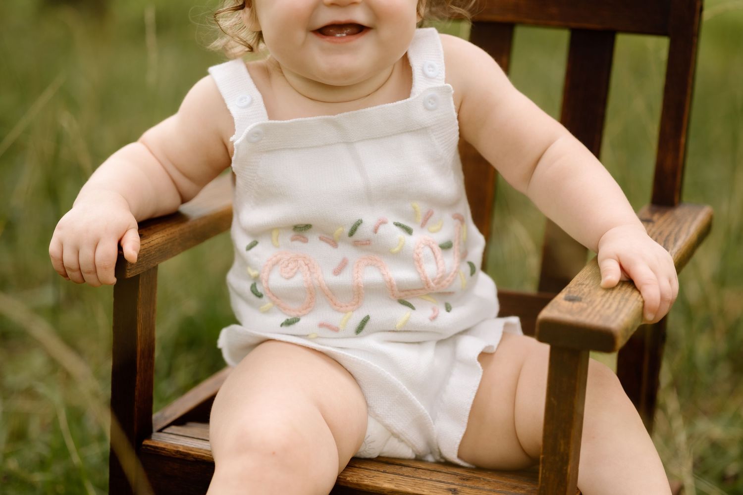 Smiling baby girl in floral white romper sitting on rustic wooden chair in grassy outdoor setting.