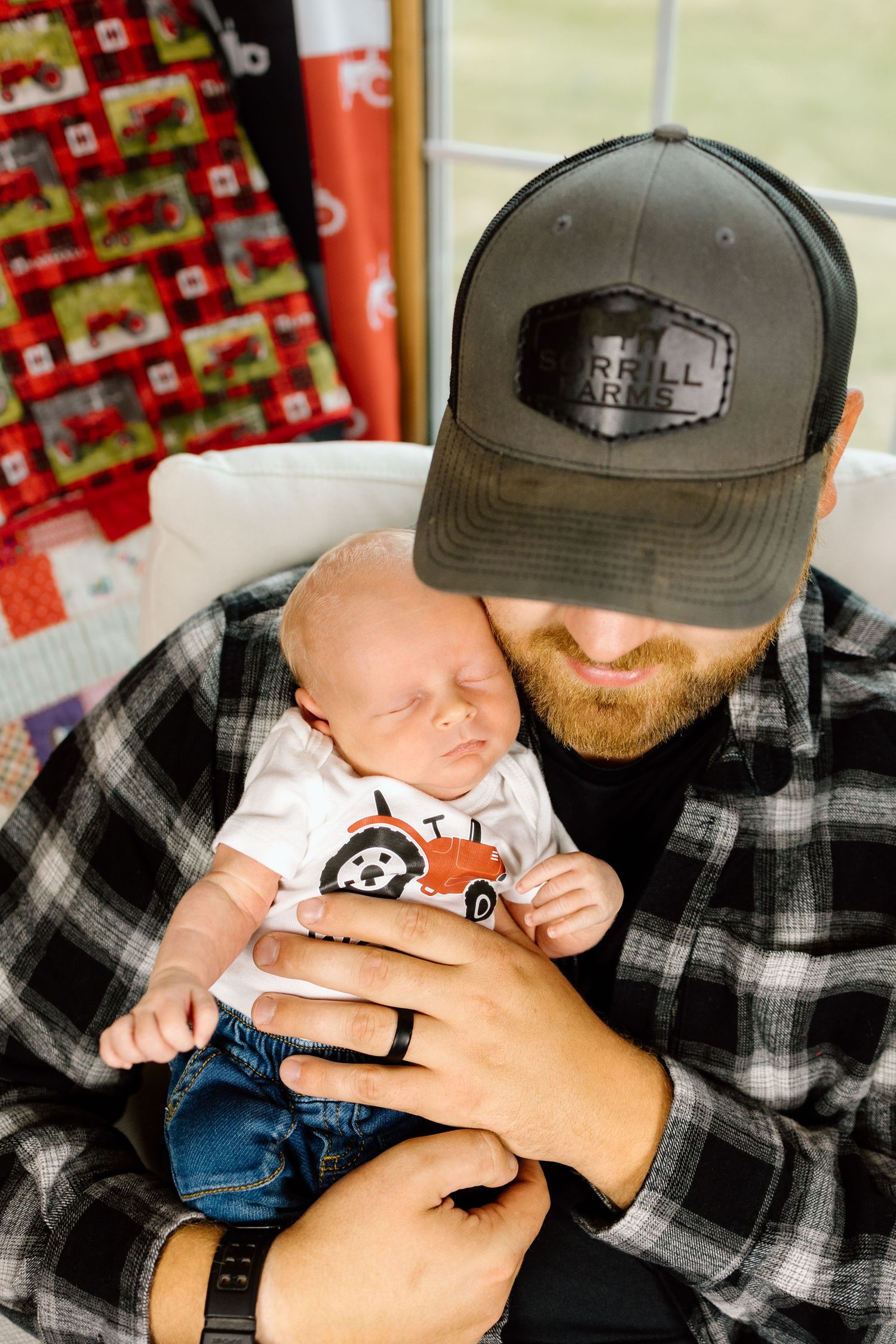 Bearded father in plaid shirt and farm cap tenderly holding his sleeping newborn baby.