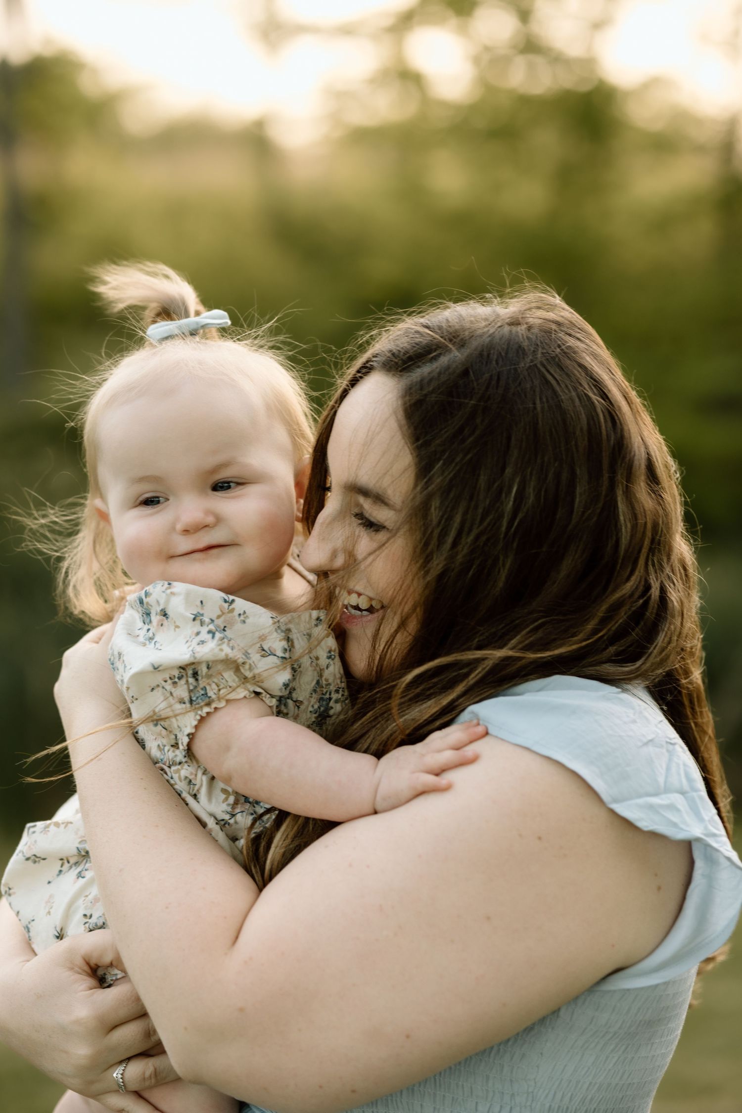 A joyful mother hugs and nuzzles her smiling baby girl outdoors in warm golden hour sunlight.