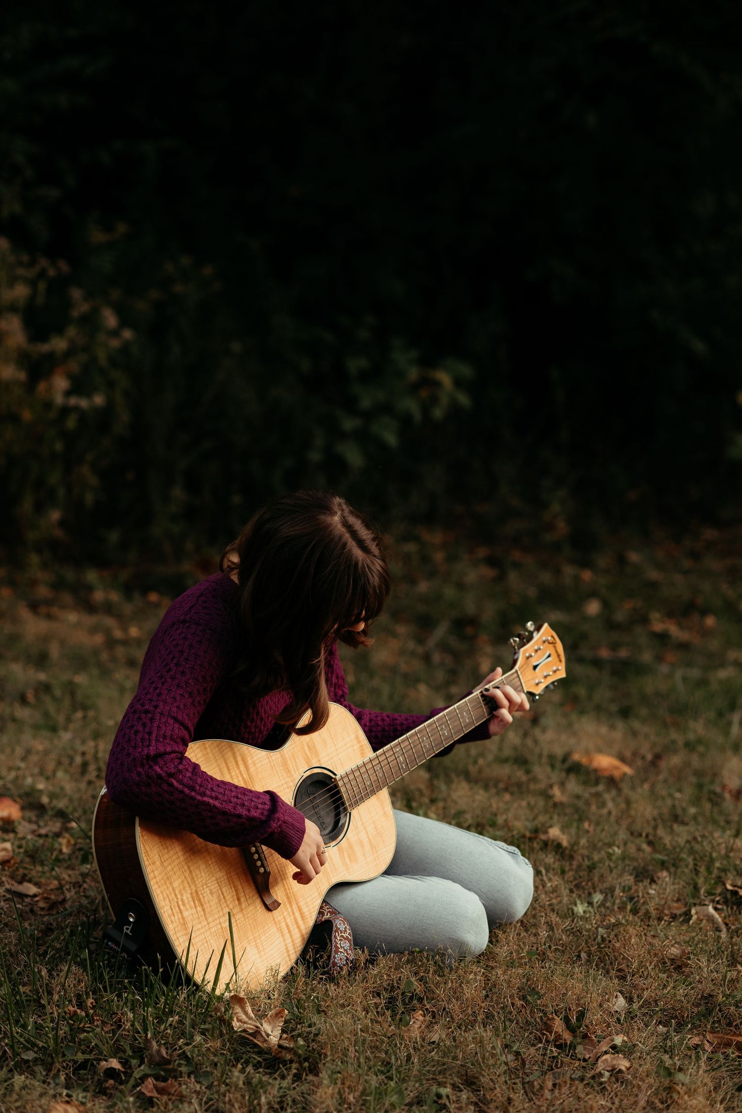 Young woman in purple sweater sits cross-legged on grass playing acoustic guitar outdoors.