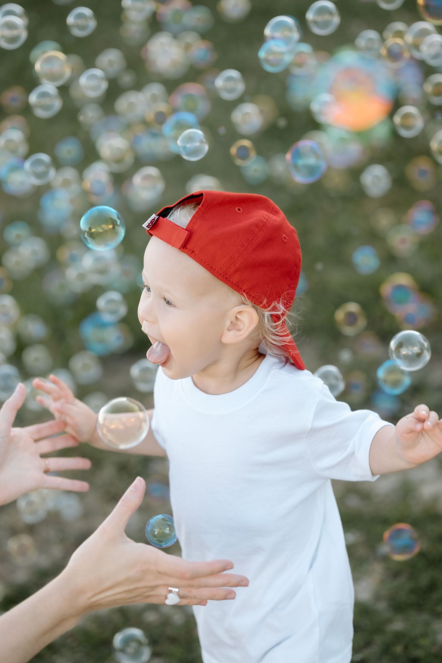 Excited toddler boy in red cap sticking tongue out trying to catch soap bubbles outside.