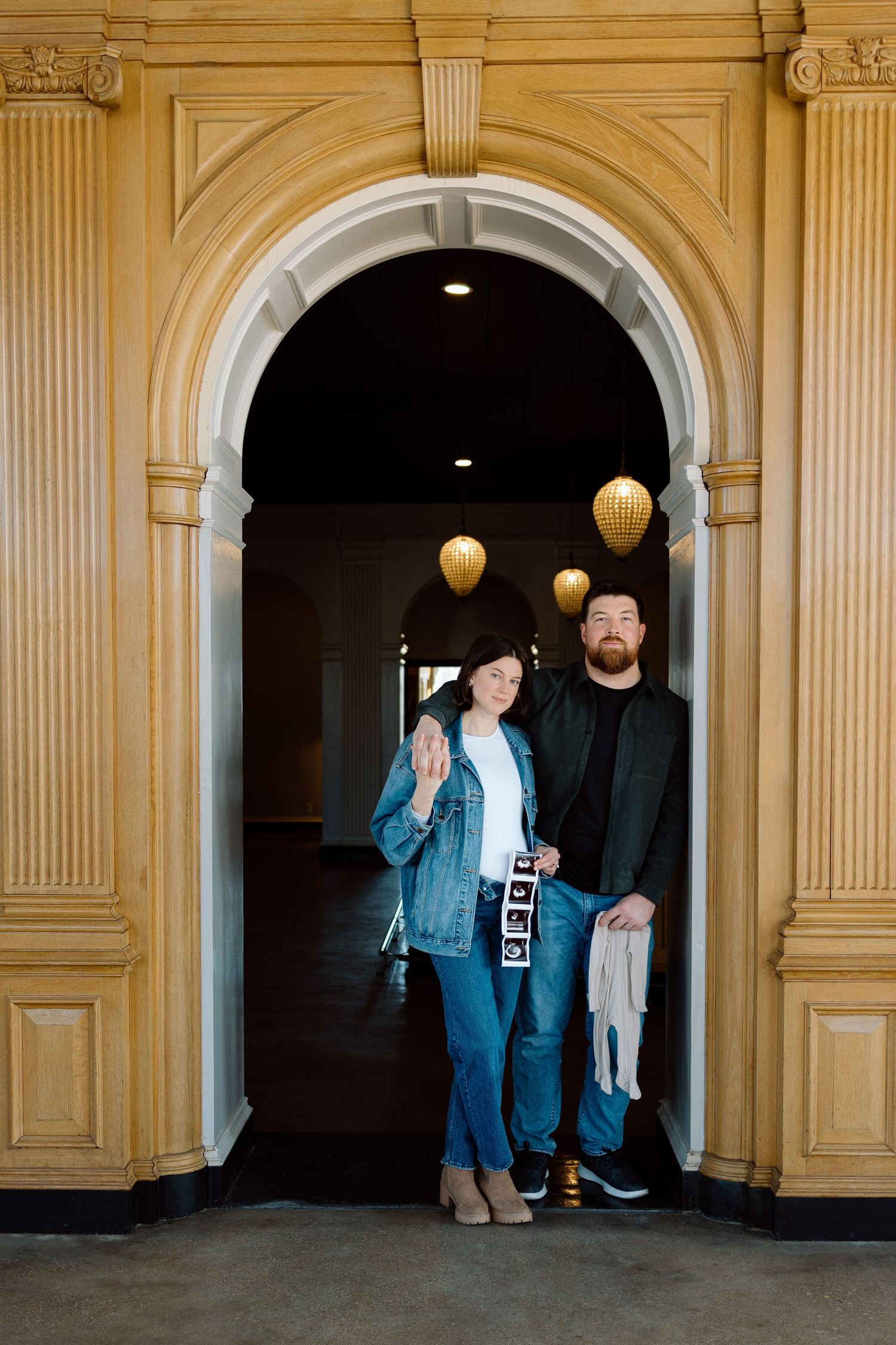 Couple stands in elegant arched doorway holding ultrasound photos and baby clothes announcing pregnancy.