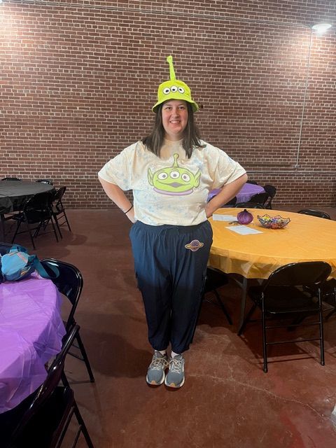 A woman poses in a Toy Story alien costume with a green three-eyed alien hat and matching graphic tee at an indoor event.