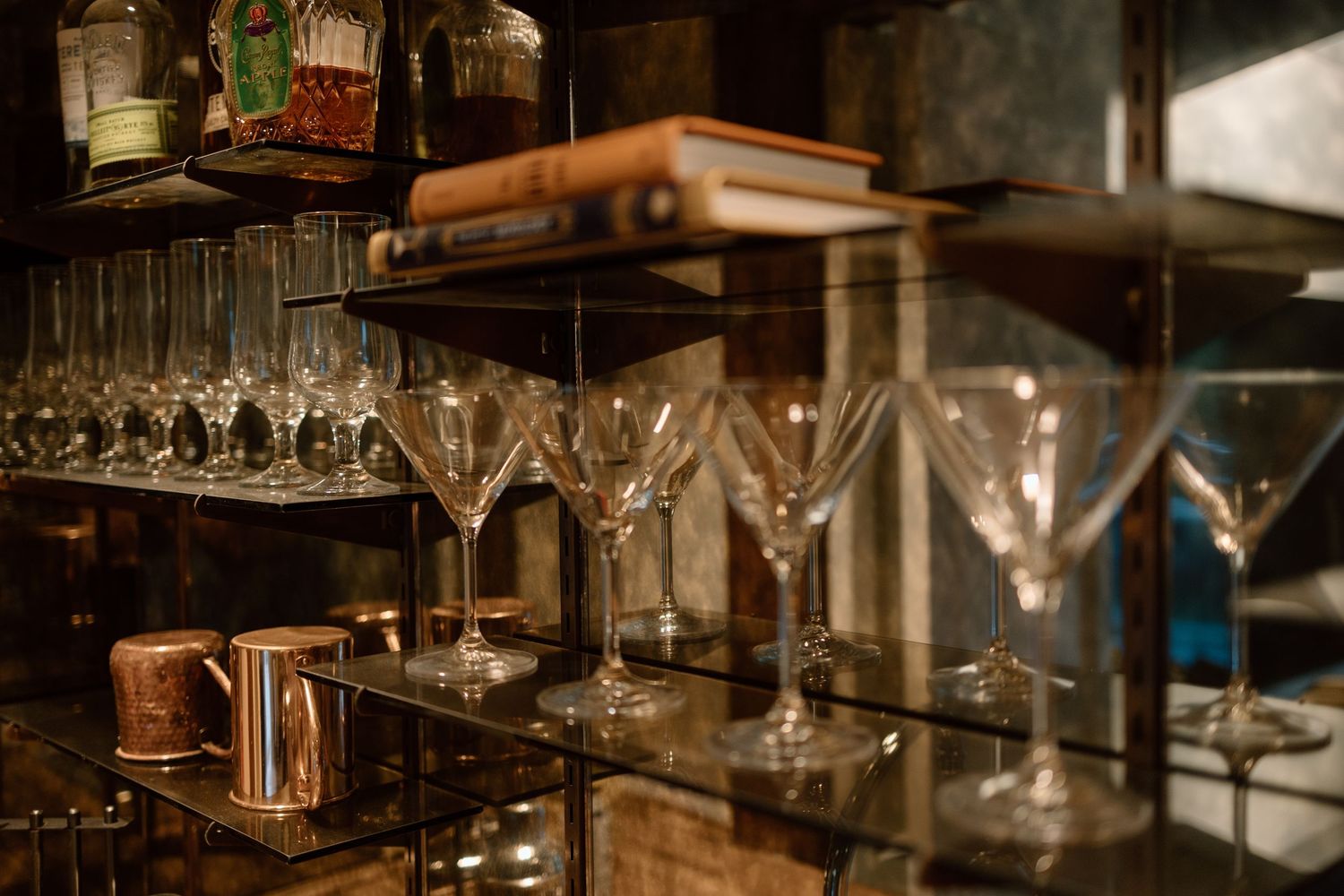 Elegant bar shelves displaying crystal martini glasses and copper mugs in warm ambient lighting.