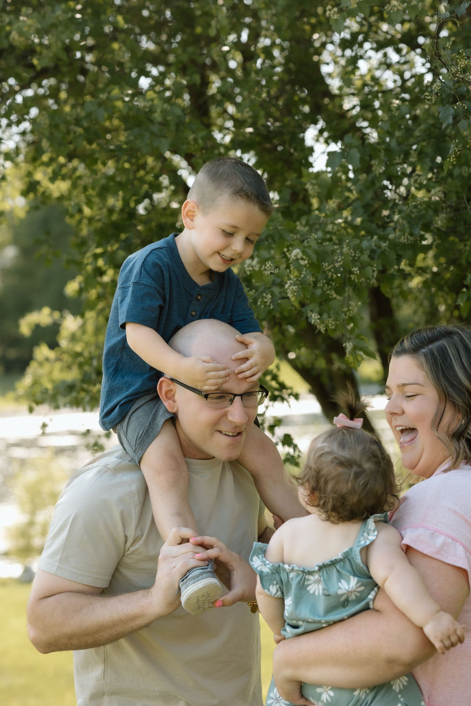 Laughing family of four enjoys playful outdoor portraits with son on dad's shoulders and baby girl in mom's arms.