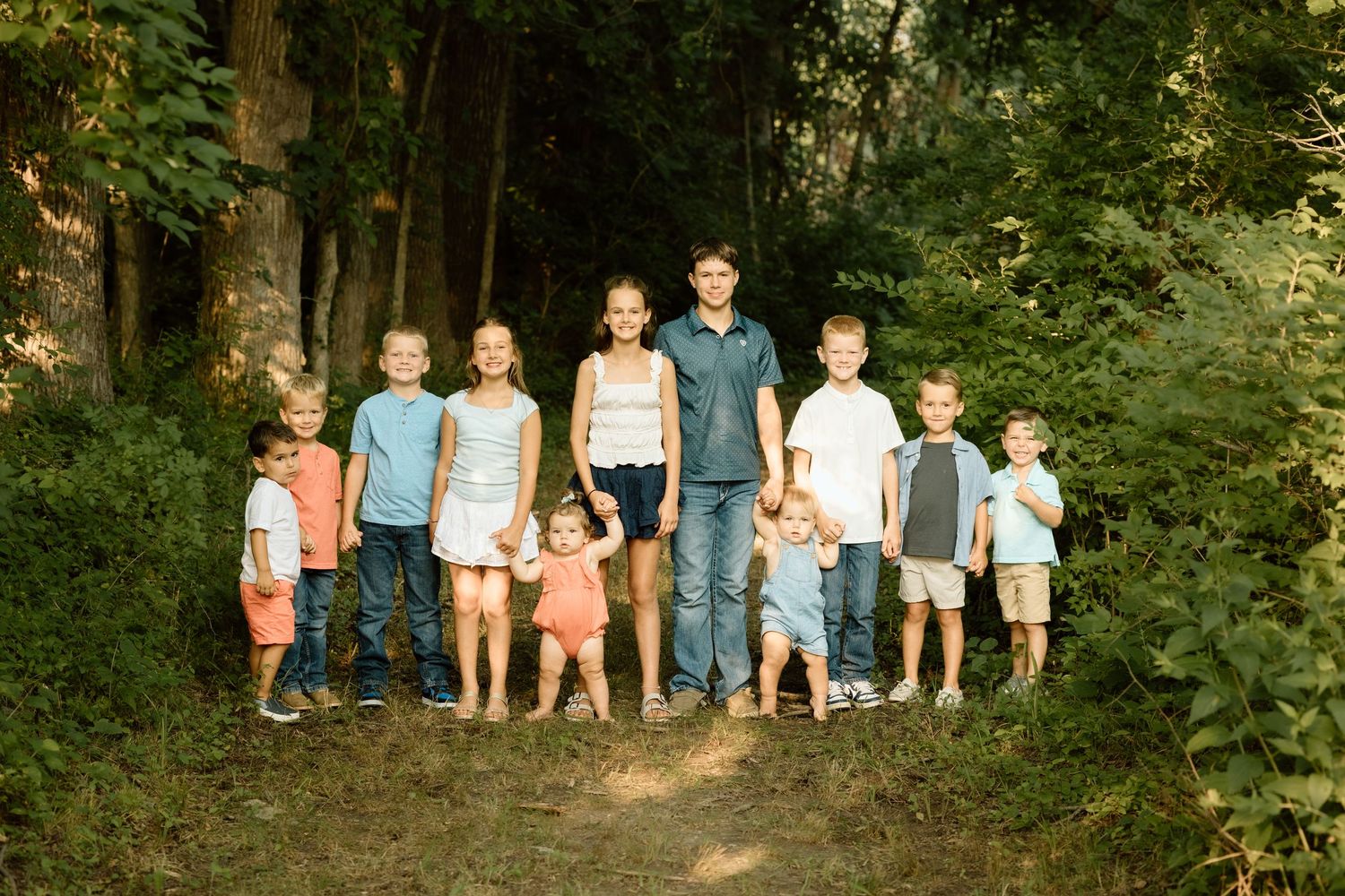 Large group of children stand together on a dirt path surrounded by green trees for an outdoor family photo.