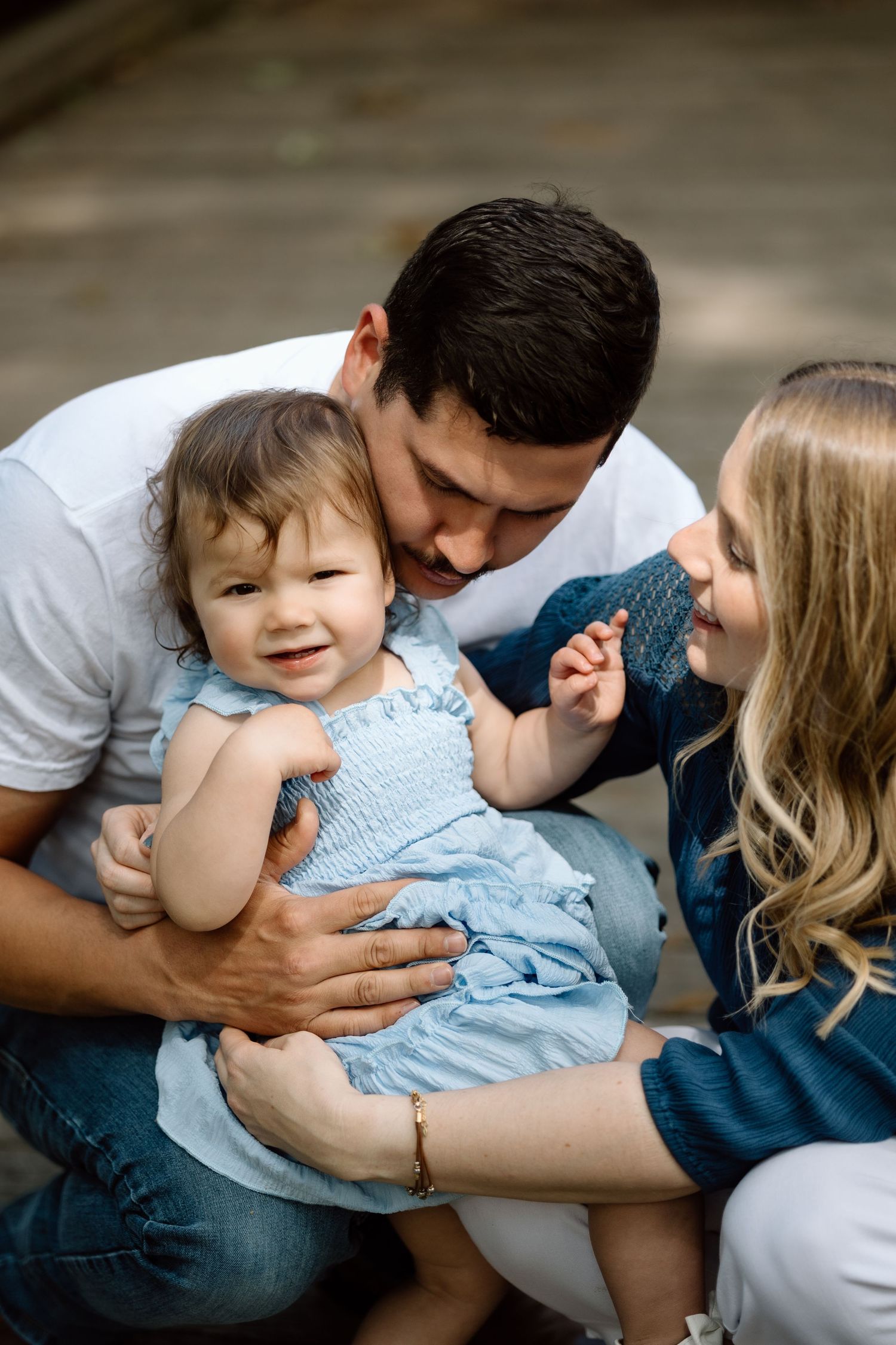 Parents lovingly kiss and cuddle their smiling toddler daughter during an outdoor family portrait session.