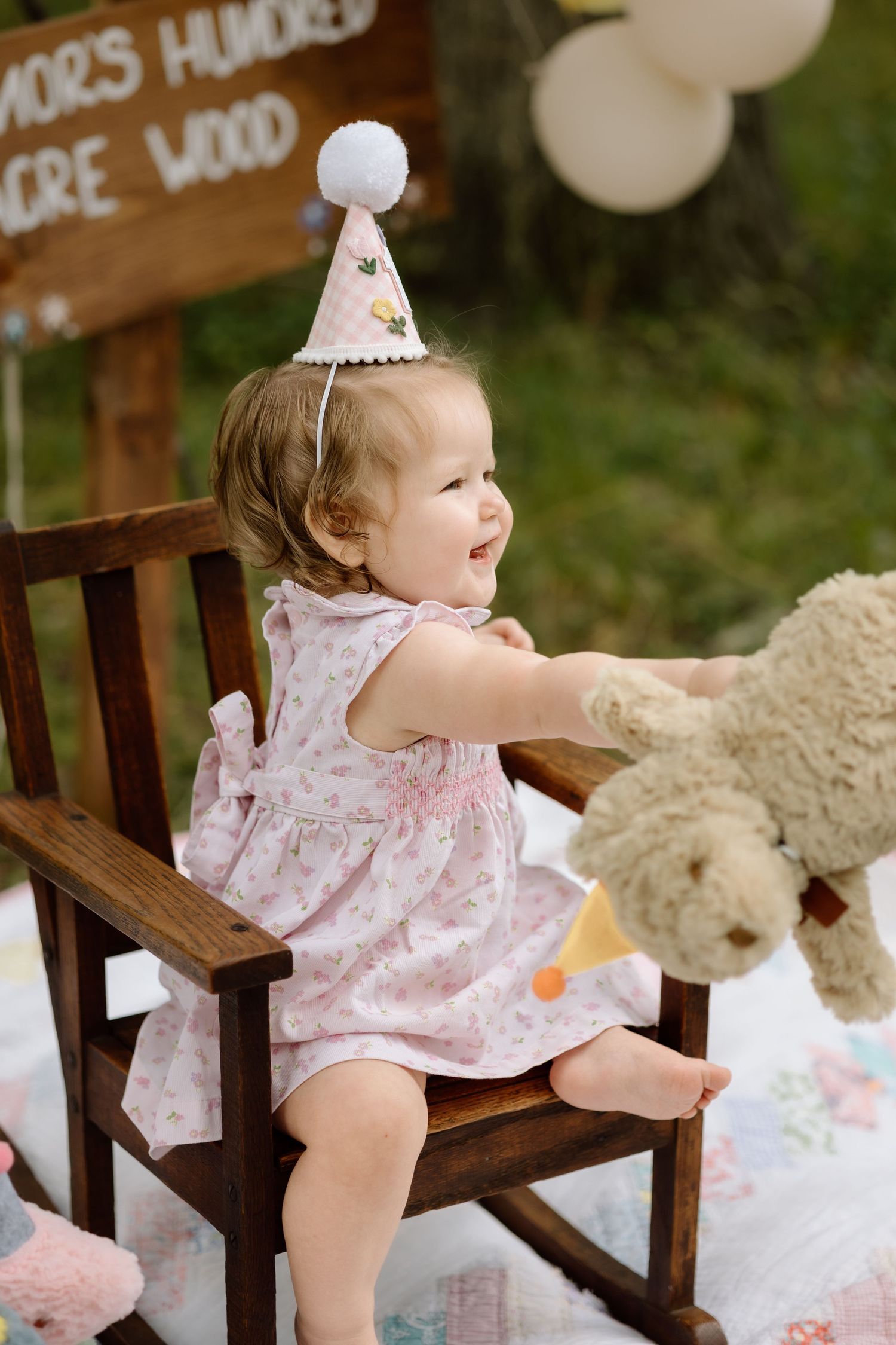 Joyful baby girl in floral dress and birthday hat hugging teddy bear outside.