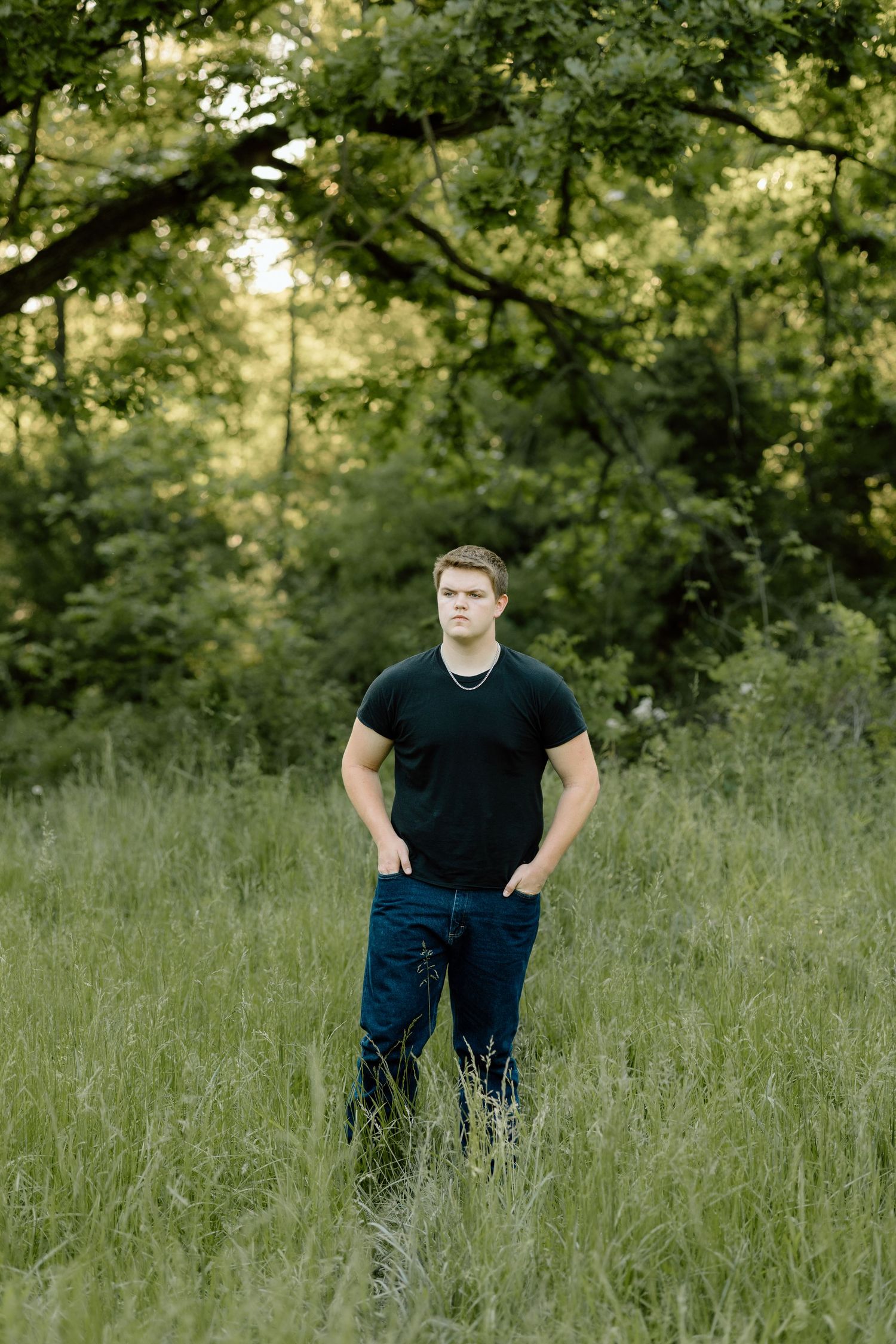 Teen boy in black t-shirt stands in tall grass beneath green trees in natural setting.