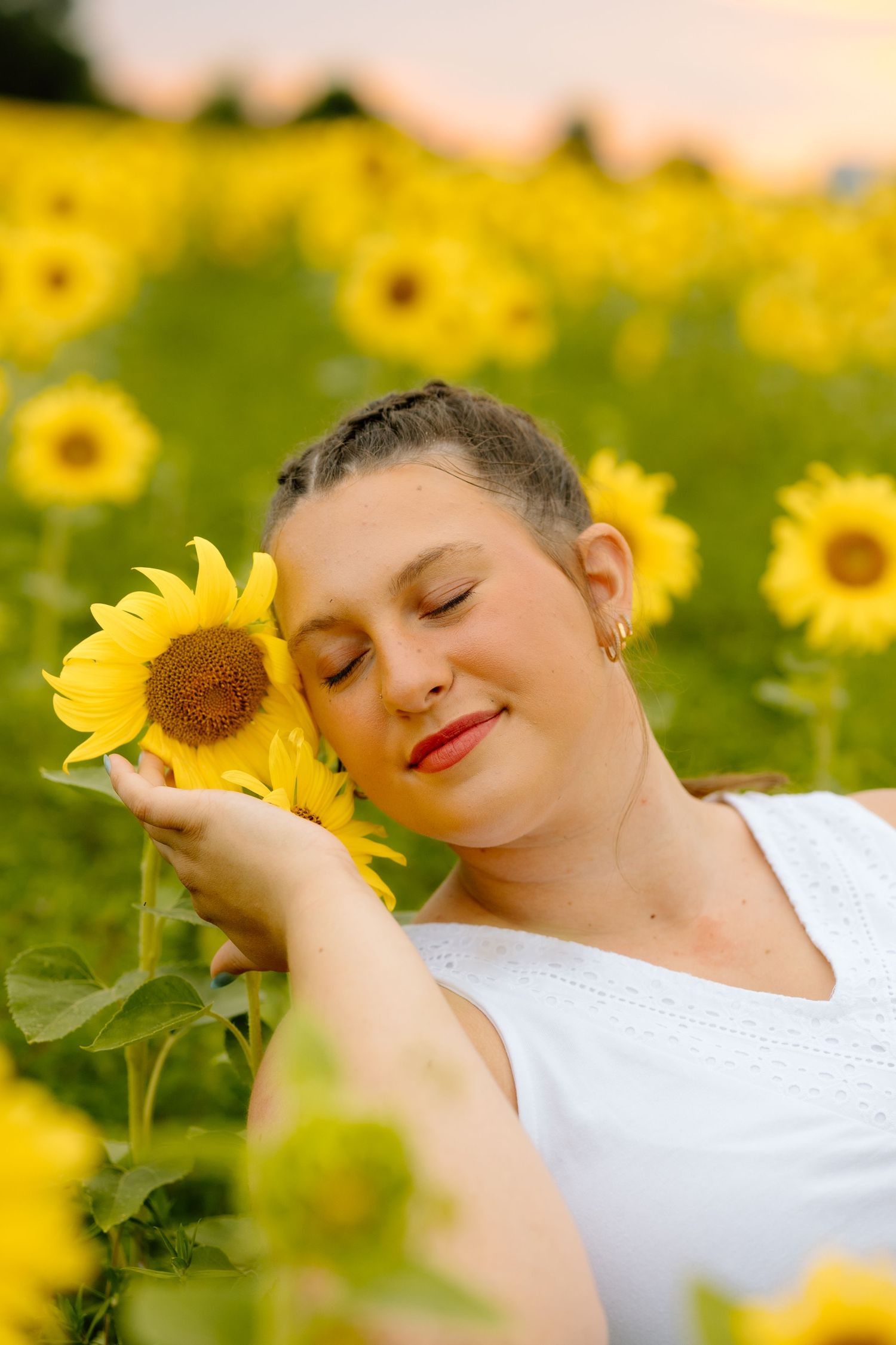 Woman in white dress eyes closed holding a sunflower to her cheek in a sunflower field.