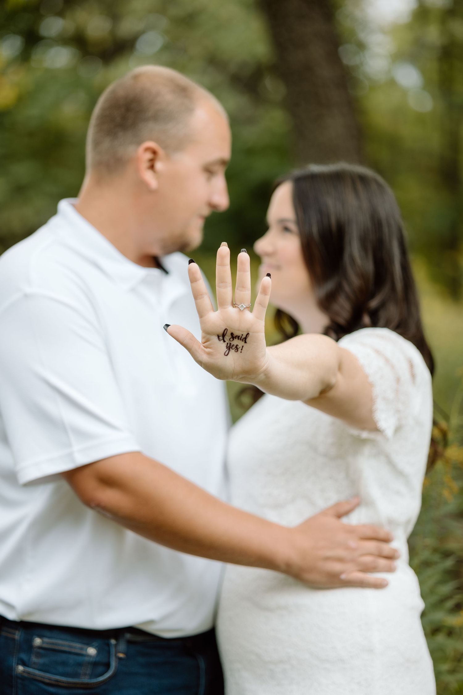 Engaged couple outdoors, woman showing palm with 'I said yes' written on her hand.