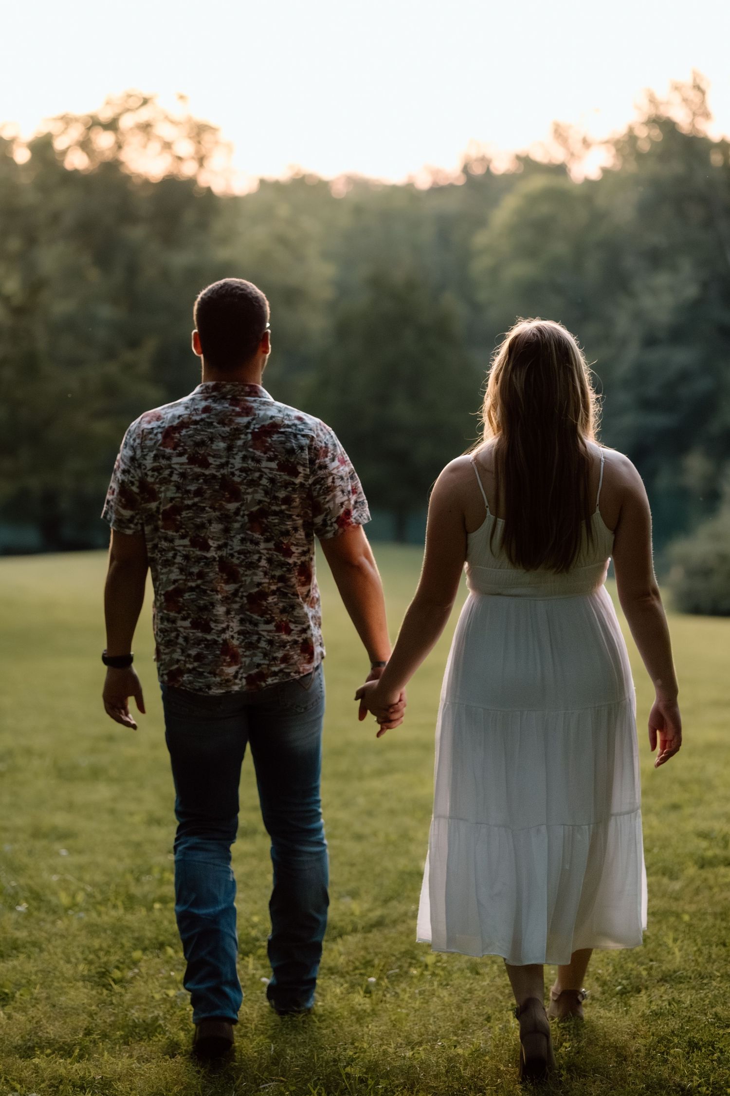 Couple holding hands walking through green field at golden hour sunset from behind.