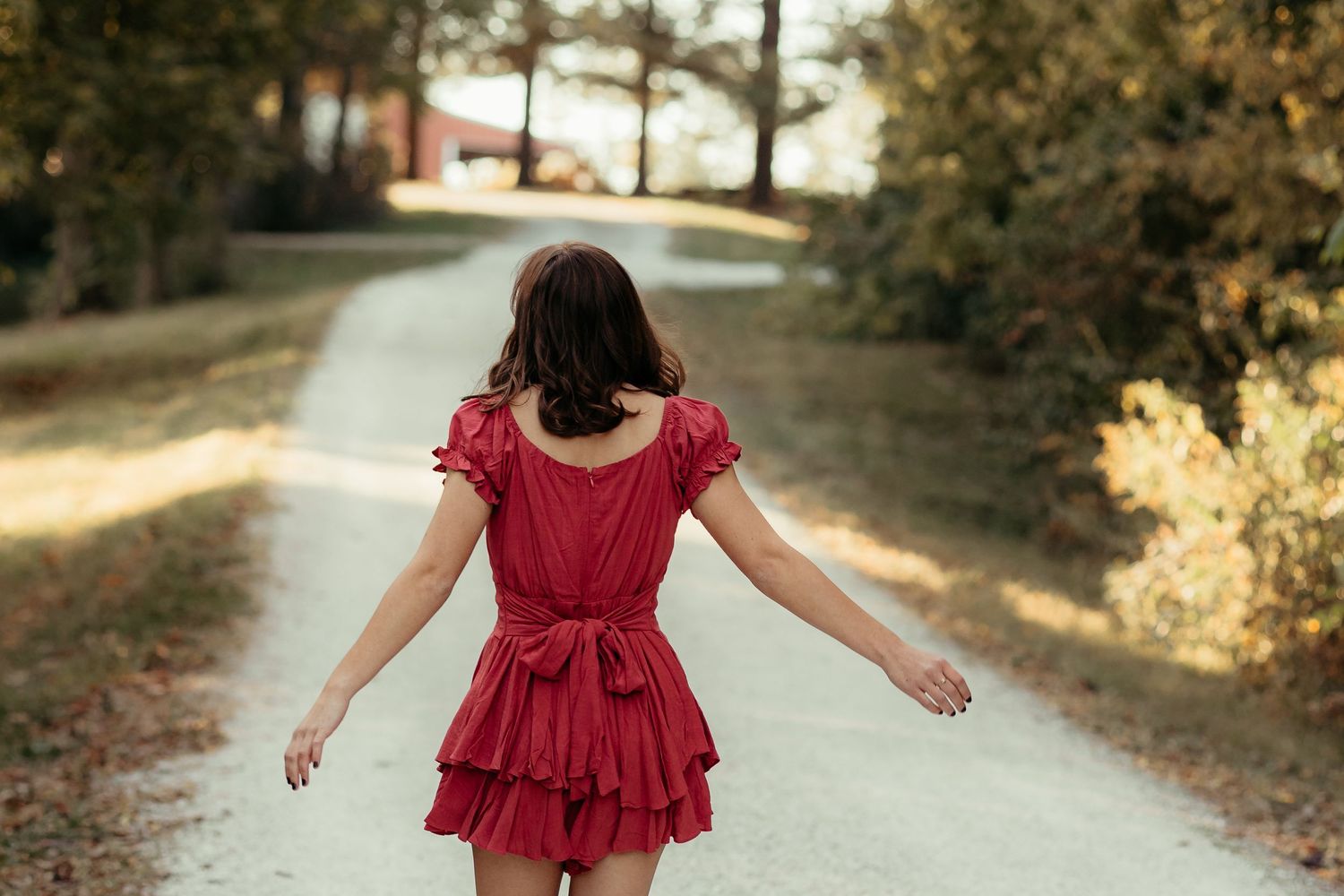 Young woman in red ruffled dress walking down a sunlit country path with arms outstretched.
