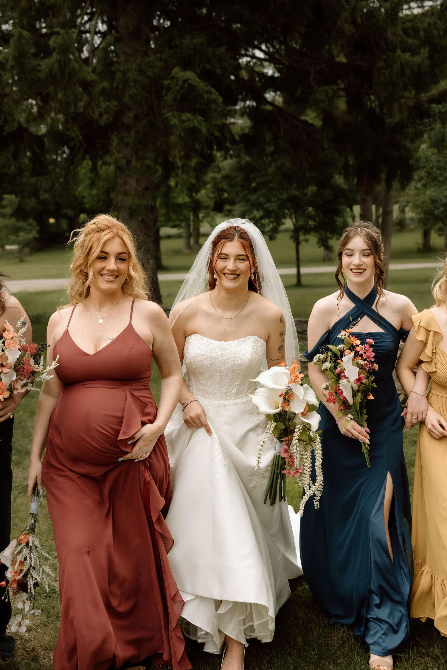 Bride in white strapless gown walks with bridesmaids in rust and teal dresses holding bouquets.