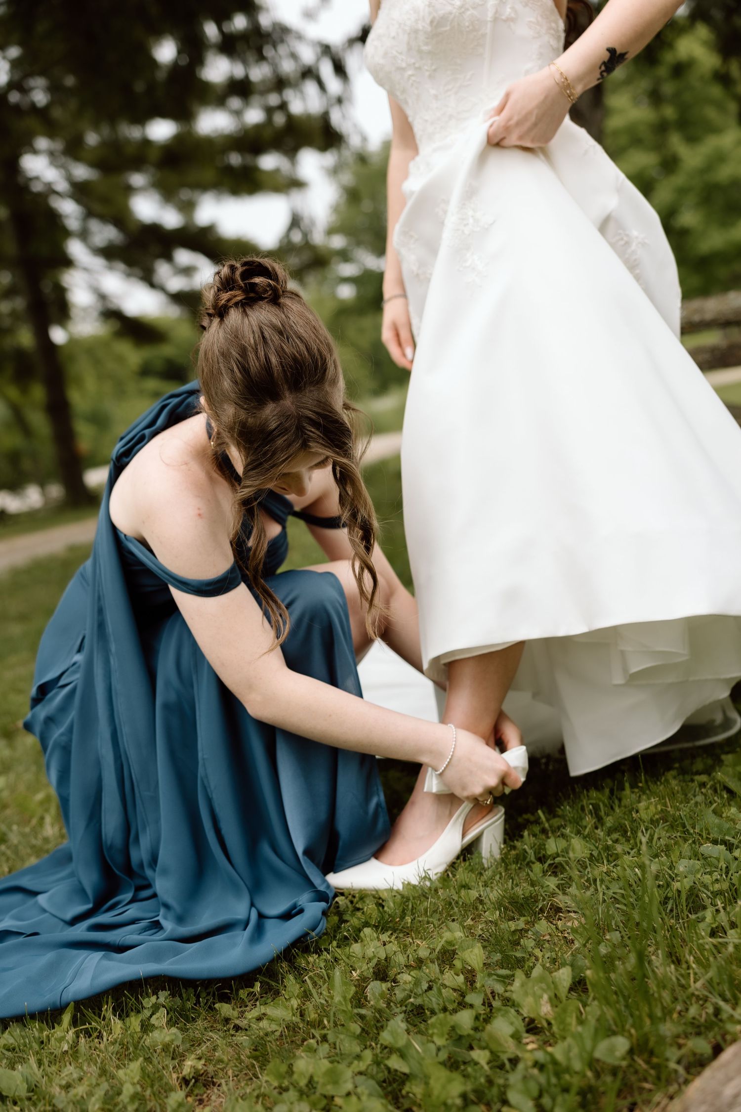 Bridesmaid in blue dress kneeling to help bride with white heels on grass outdoors.