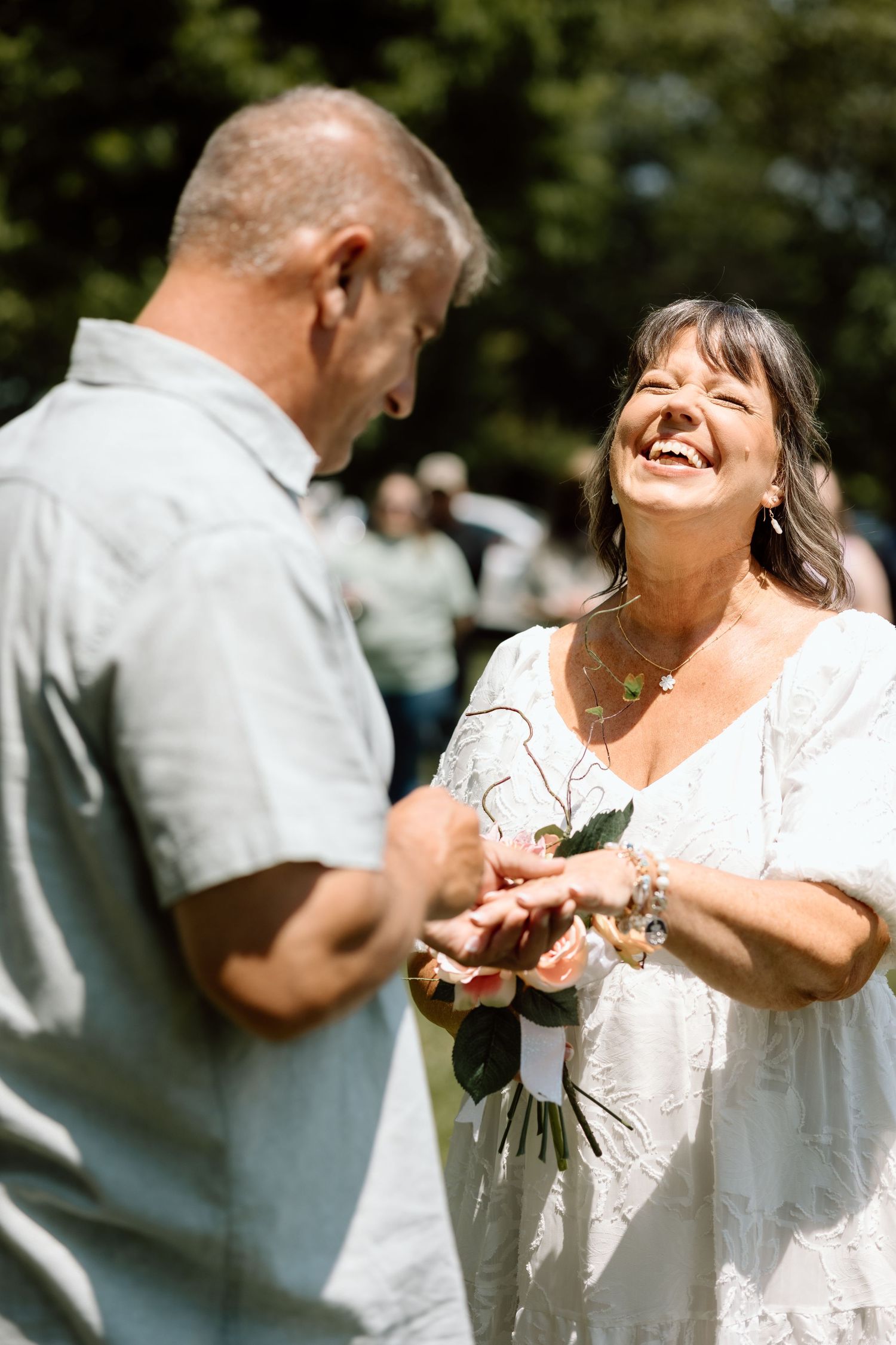 Bride laughs joyfully as groom places wedding ring on her finger during outdoor ceremony.