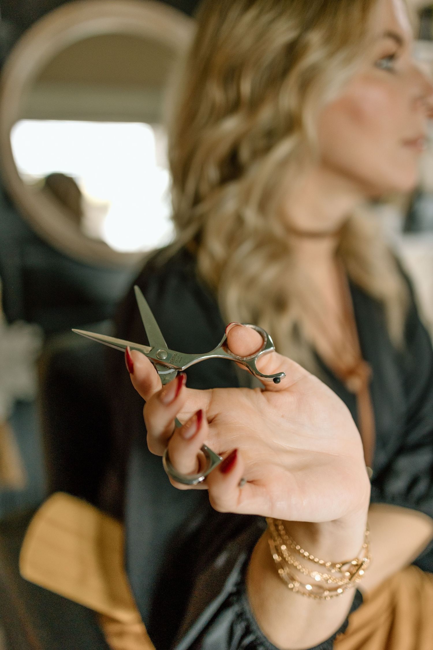Blonde female hairstylist holding silver scissors with red manicured nails and gold bracelets in a salon.