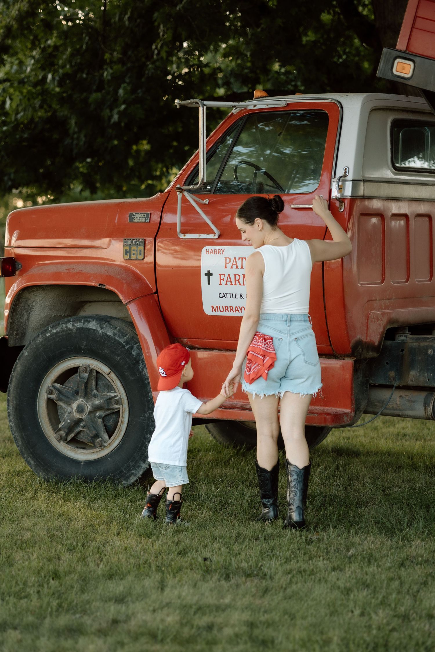 Mother and toddler son holding hands beside a vintage red farm truck outdoors.