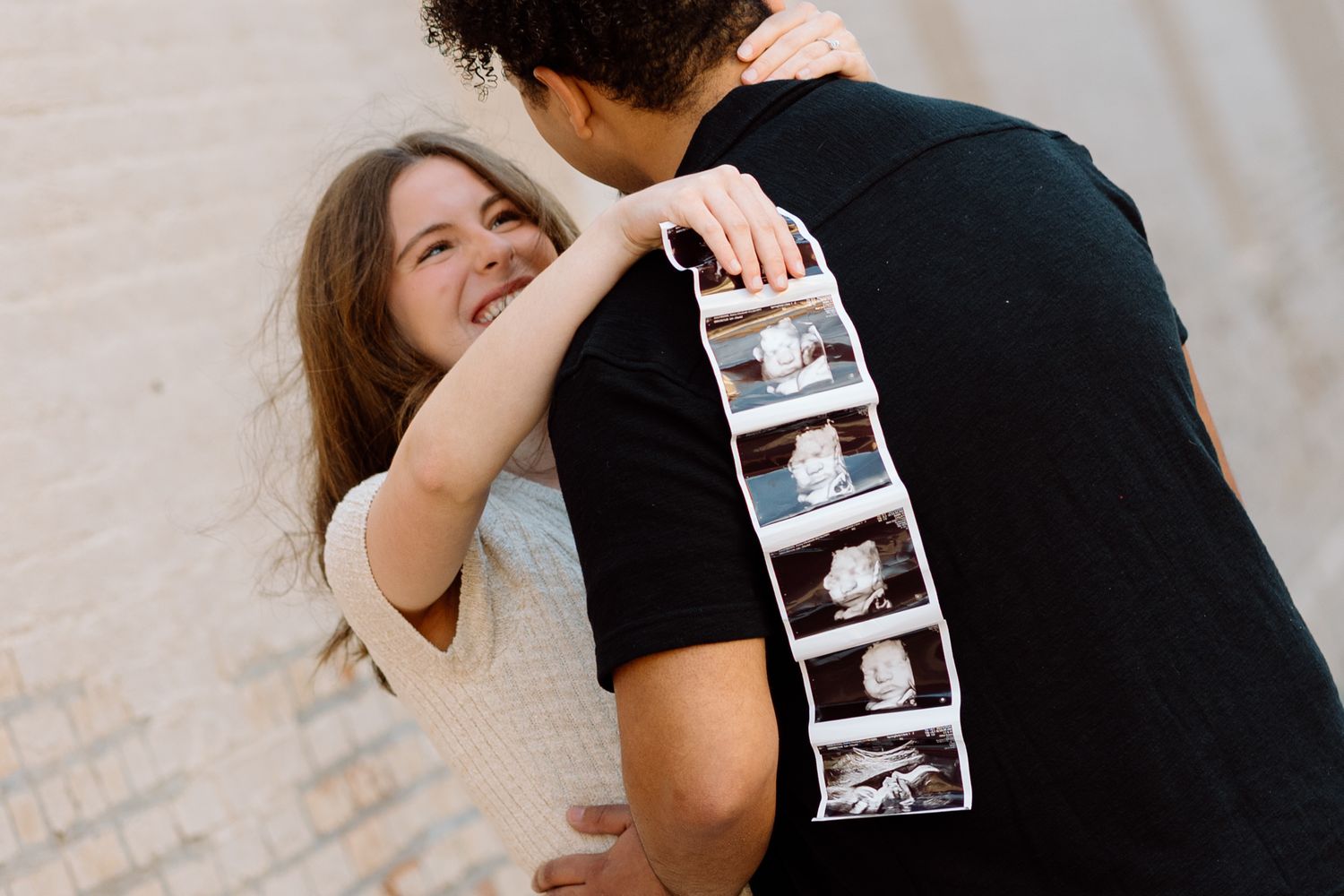Smiling pregnant woman hugging partner who holds ultrasound photo strip during outdoor maternity session.