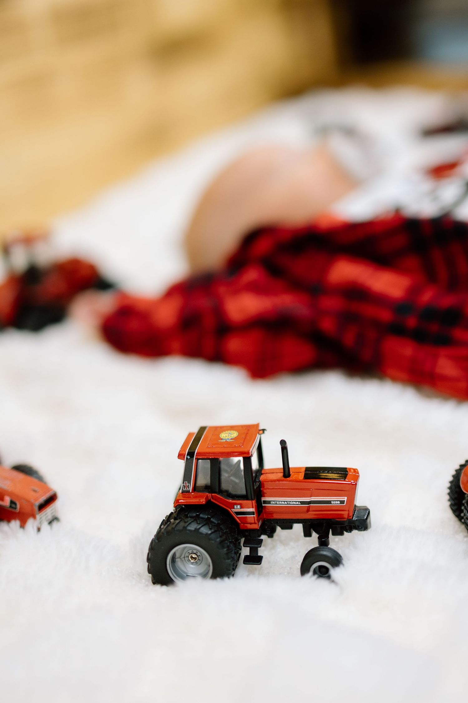 Close-up of orange toy tractor on white fluffy surface with baby in red plaid flannel shirt in background.