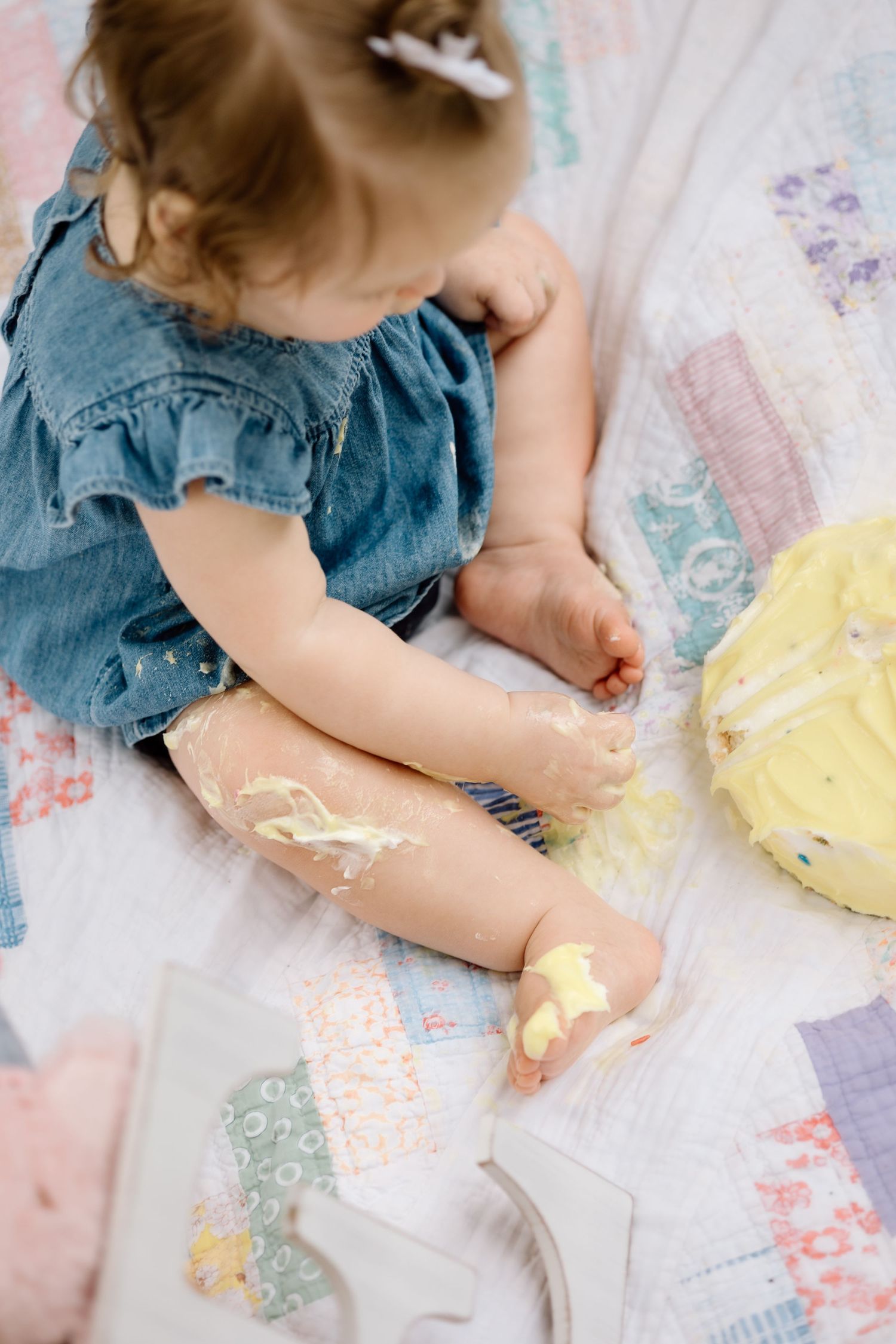 Overhead view of toddler girl in denim dress with cake smash mess on legs sitting on quilt.