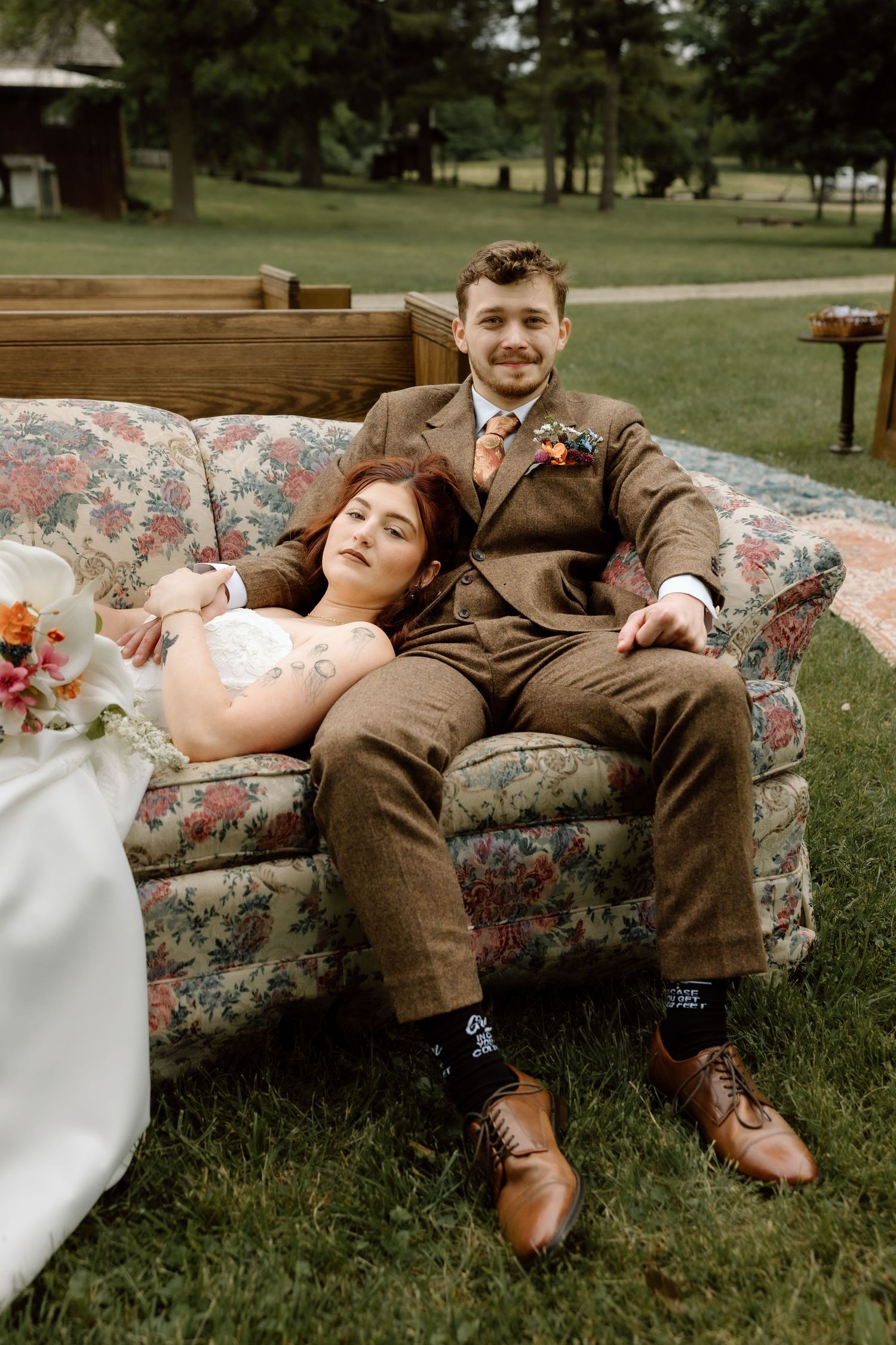 Bride and groom relaxing on a vintage floral sofa outdoors, groom in brown tweed suit smiling at camera.