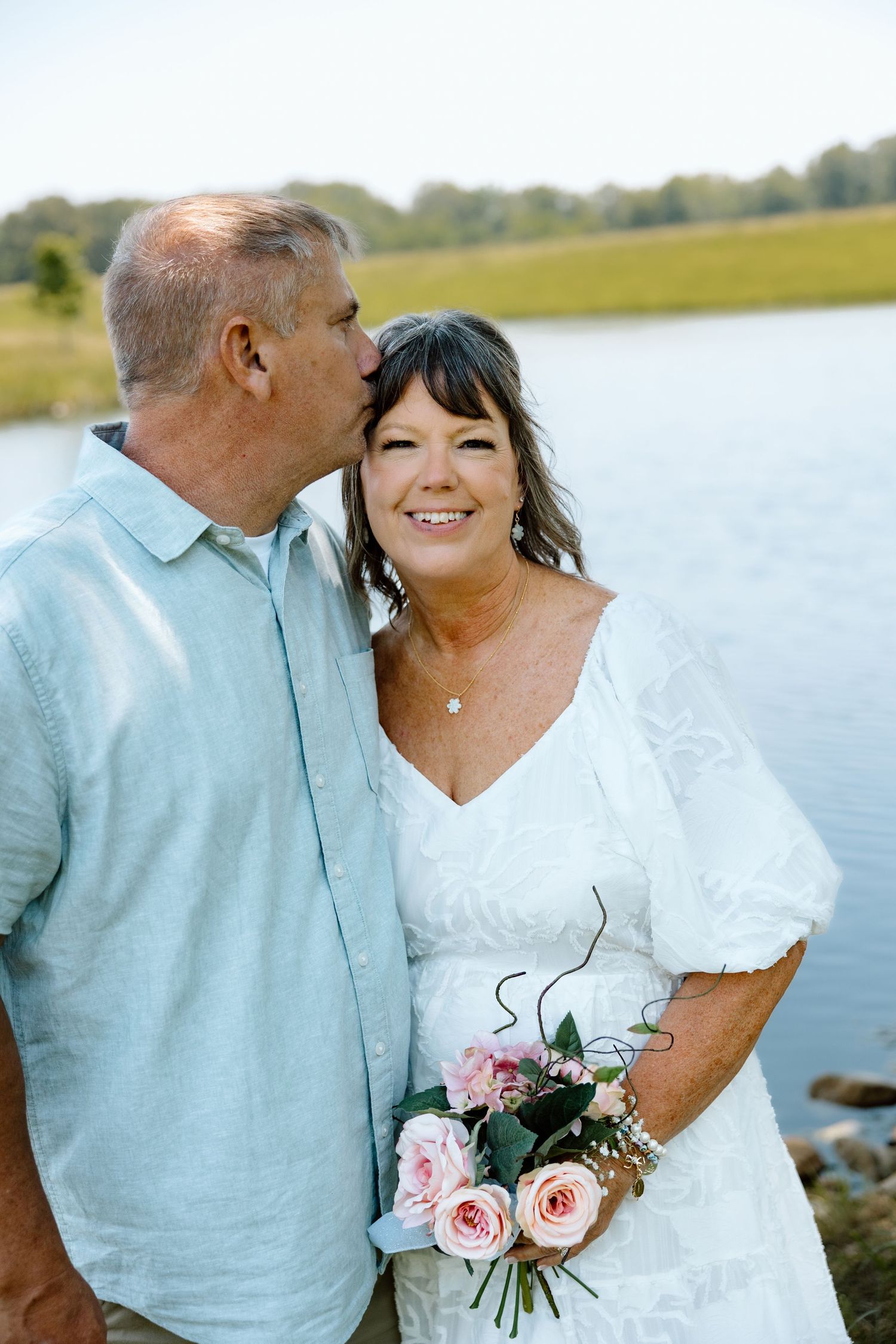 Groom kissing bride on cheek outdoors by lake, bride smiling and holding pink roses bouquet.