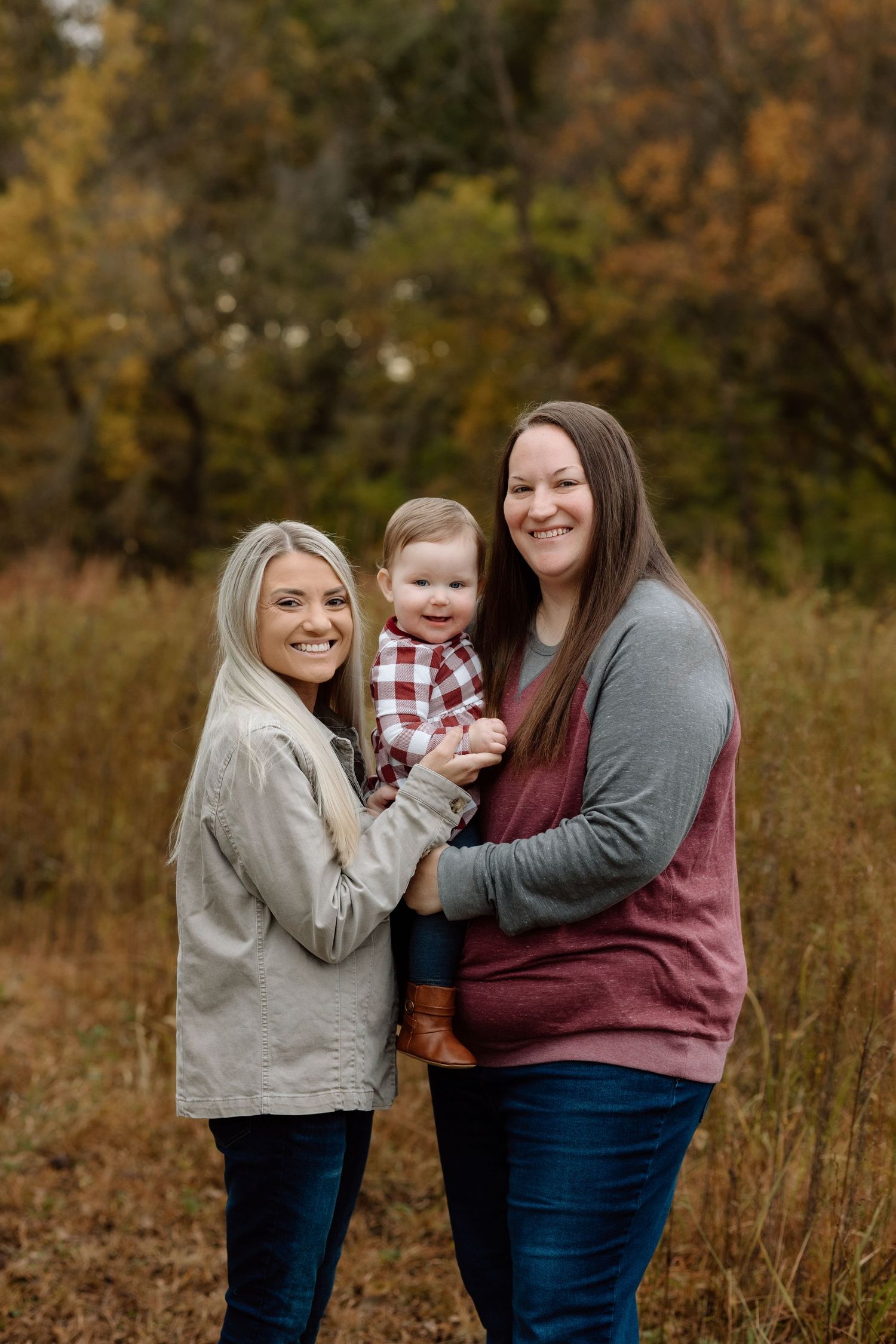 Two moms smile holding their happy toddler in a plaid outfit during fall family photos.