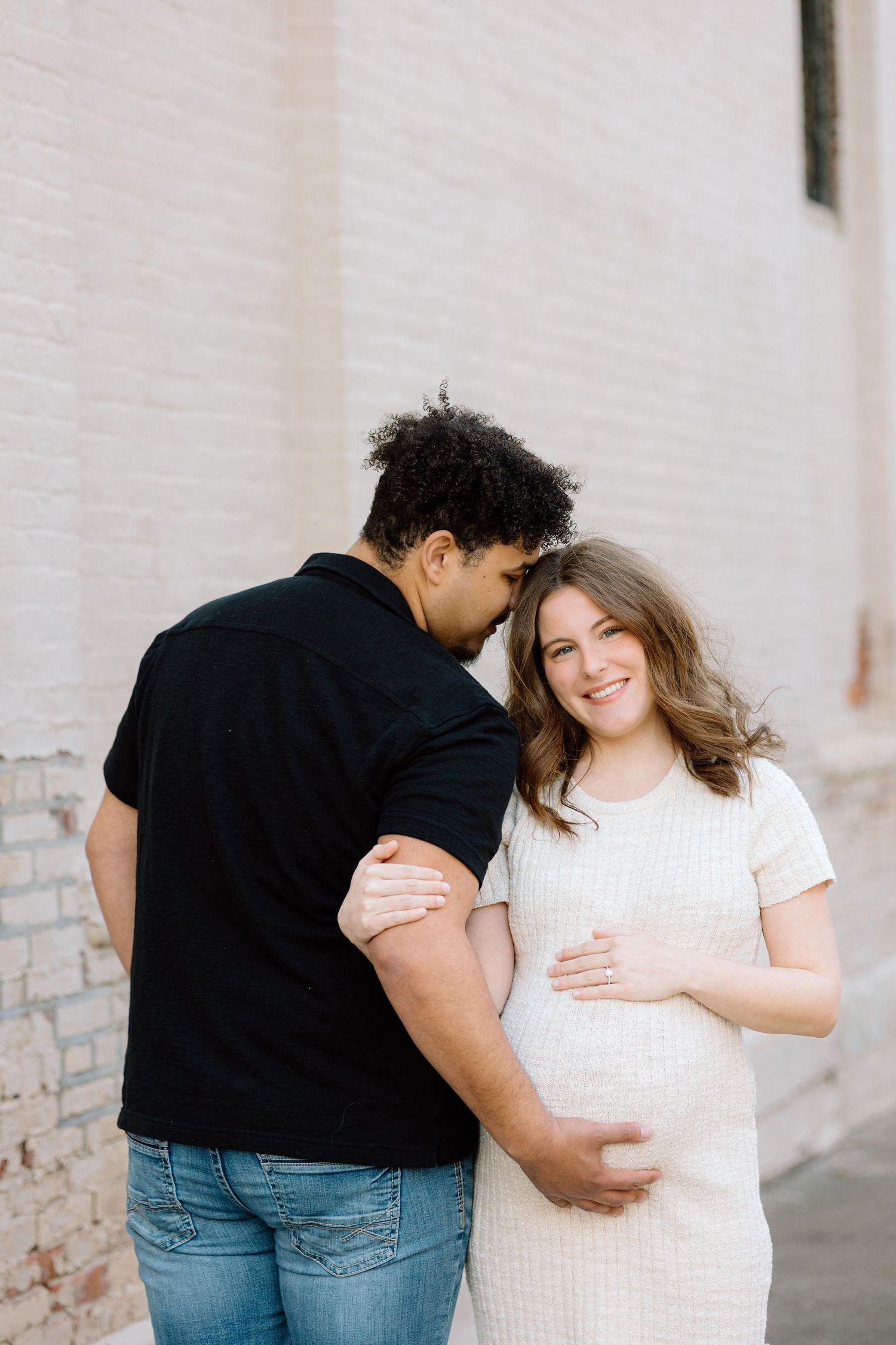 Pregnant woman in white knit dress smiles as partner in black shirt leans in close during outdoor maternity session.