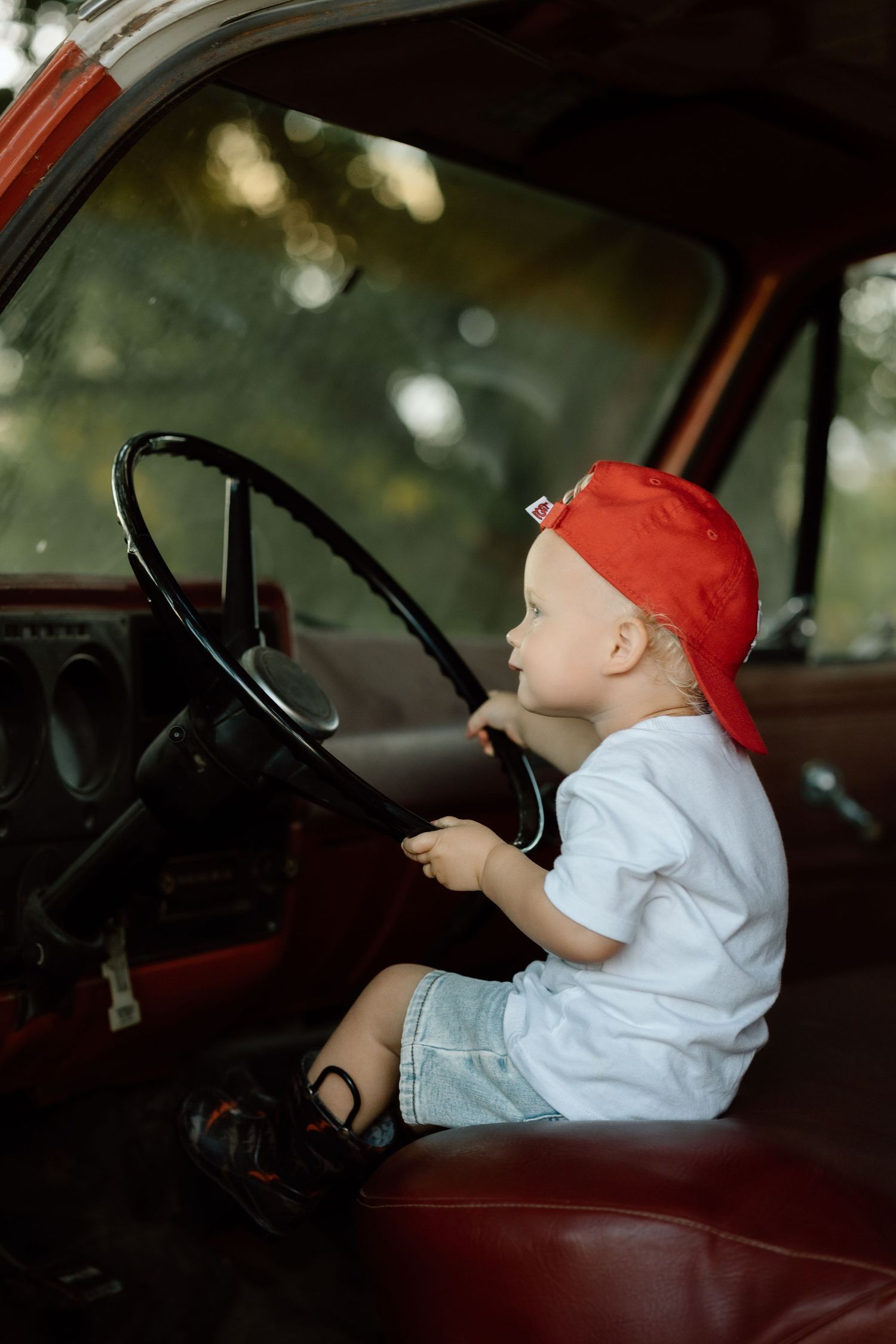 Side view of toddler in red cap pretending to drive a vintage red truck interior.