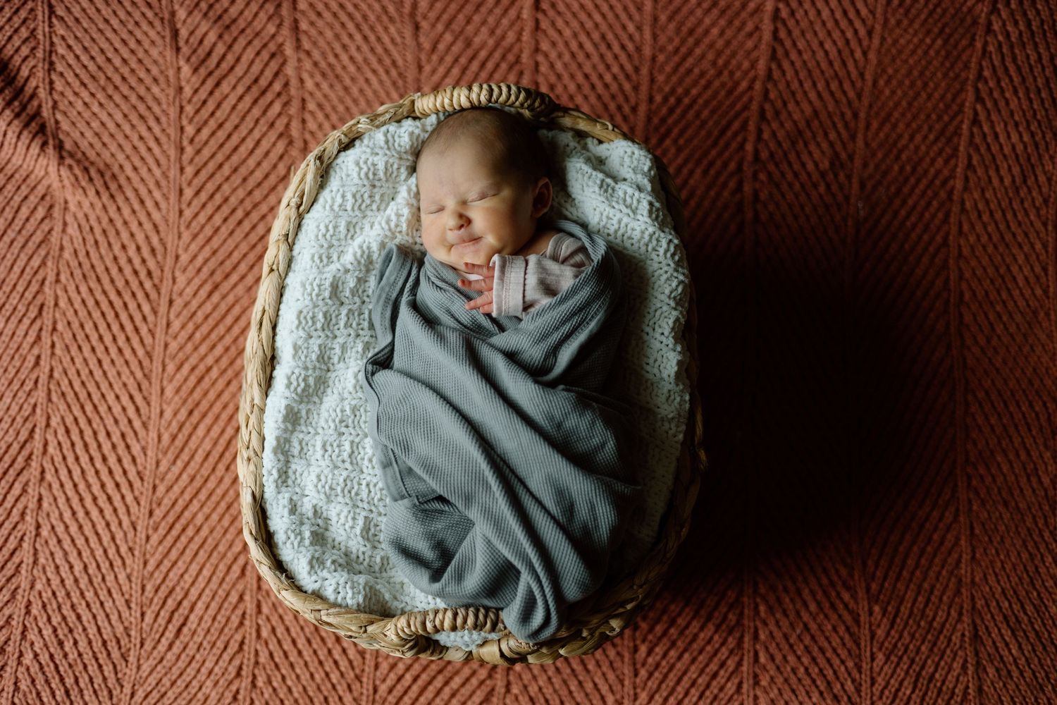 Overhead view of swaddled newborn baby sleeping peacefully in a wicker basket on rust blanket.