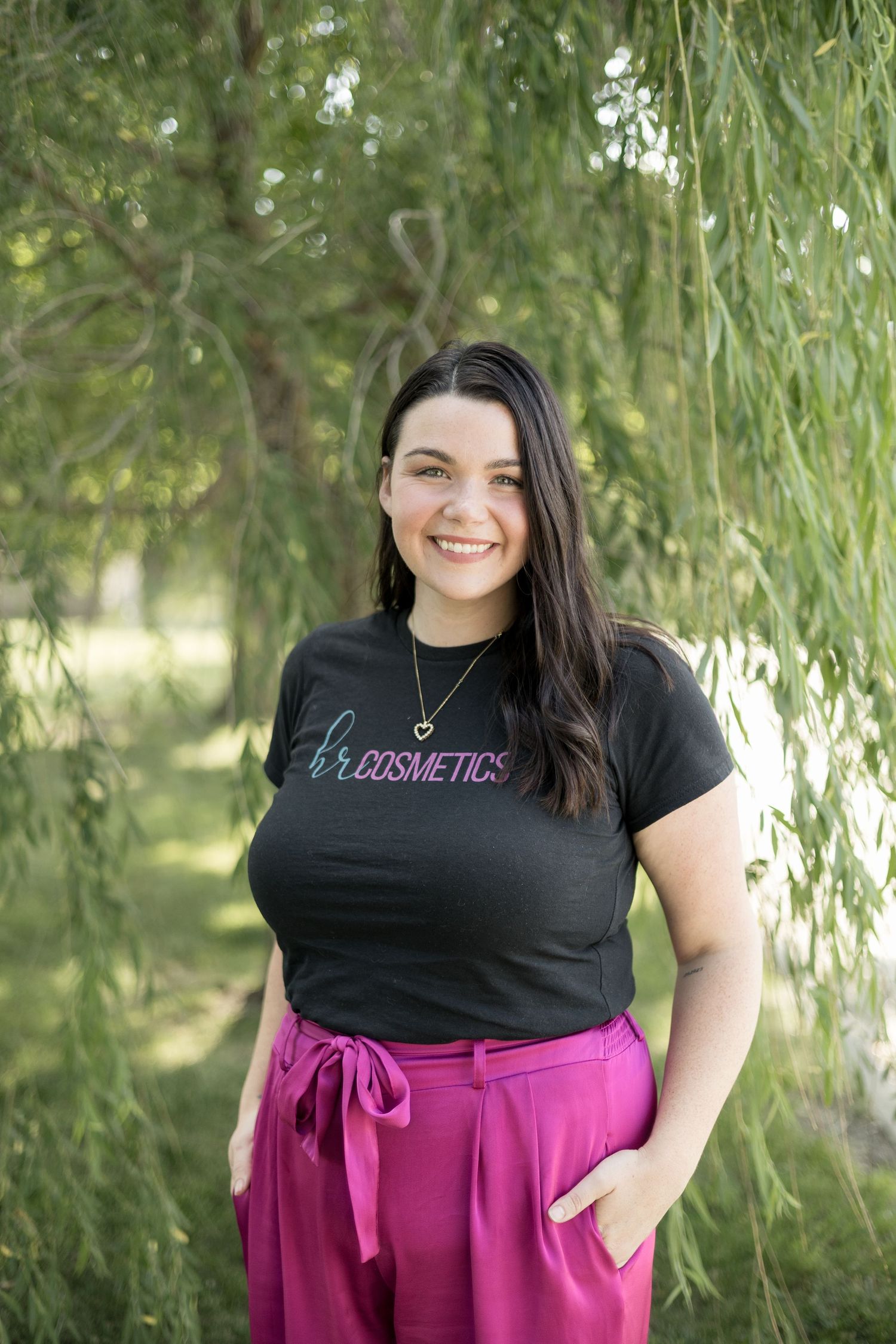 Smiling woman in HR Cosmetics black tee and pink pants posing under a weeping willow tree.
