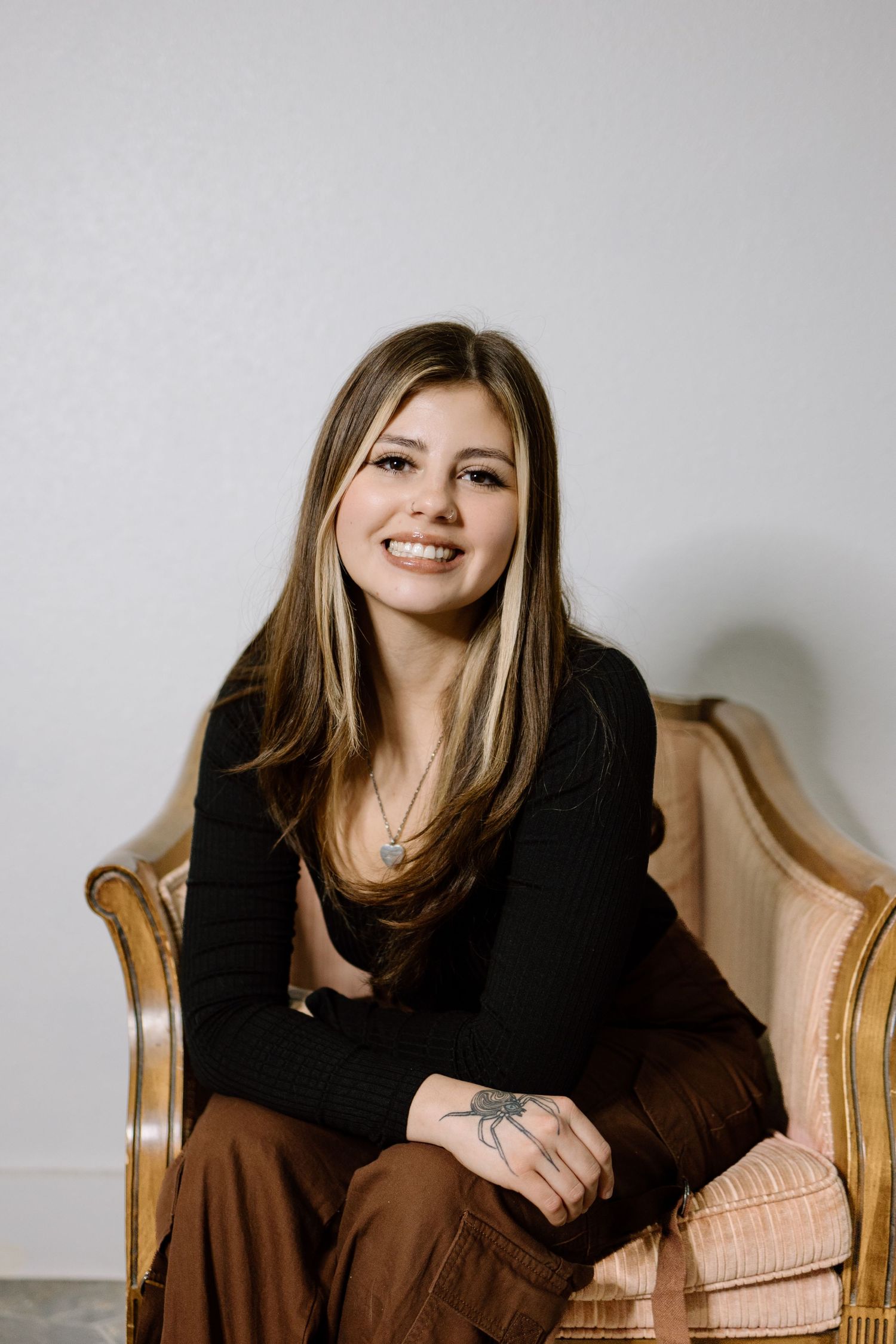 Smiling young woman with highlighted hair in black top and brown pants sitting in vintage armchair.