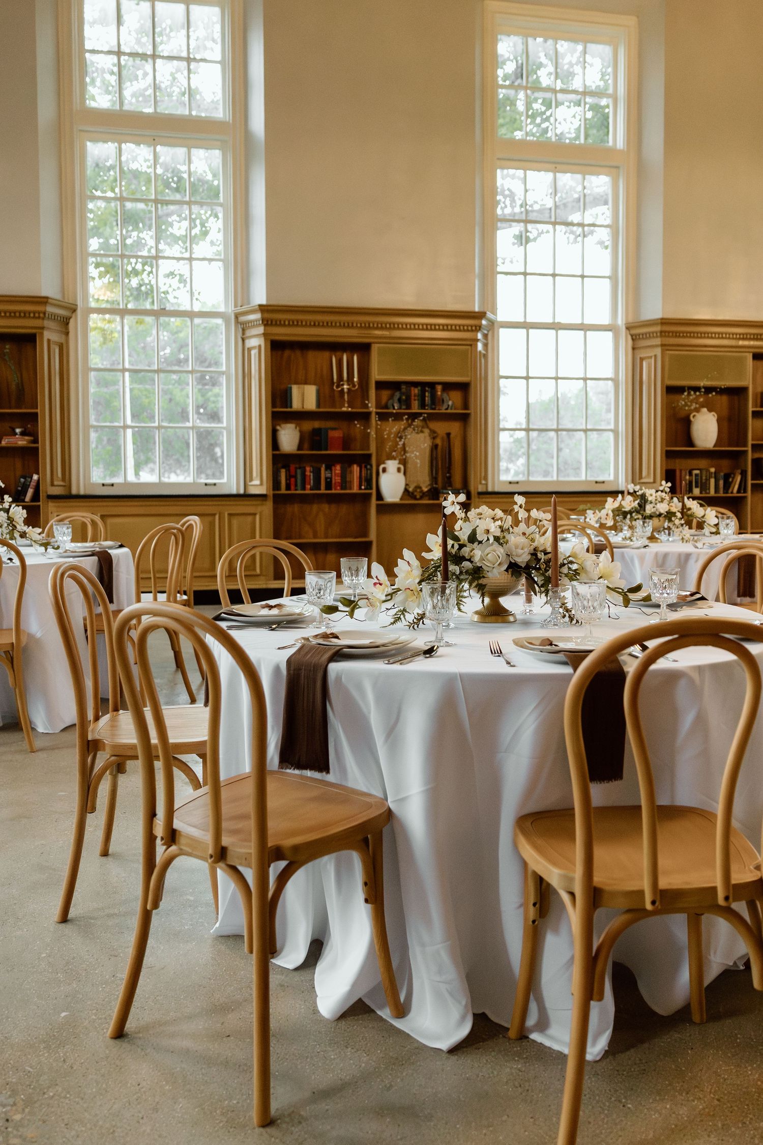 Bright wedding reception room with round tables, white linens, bentwood chairs, floral centerpieces, and tall windows.