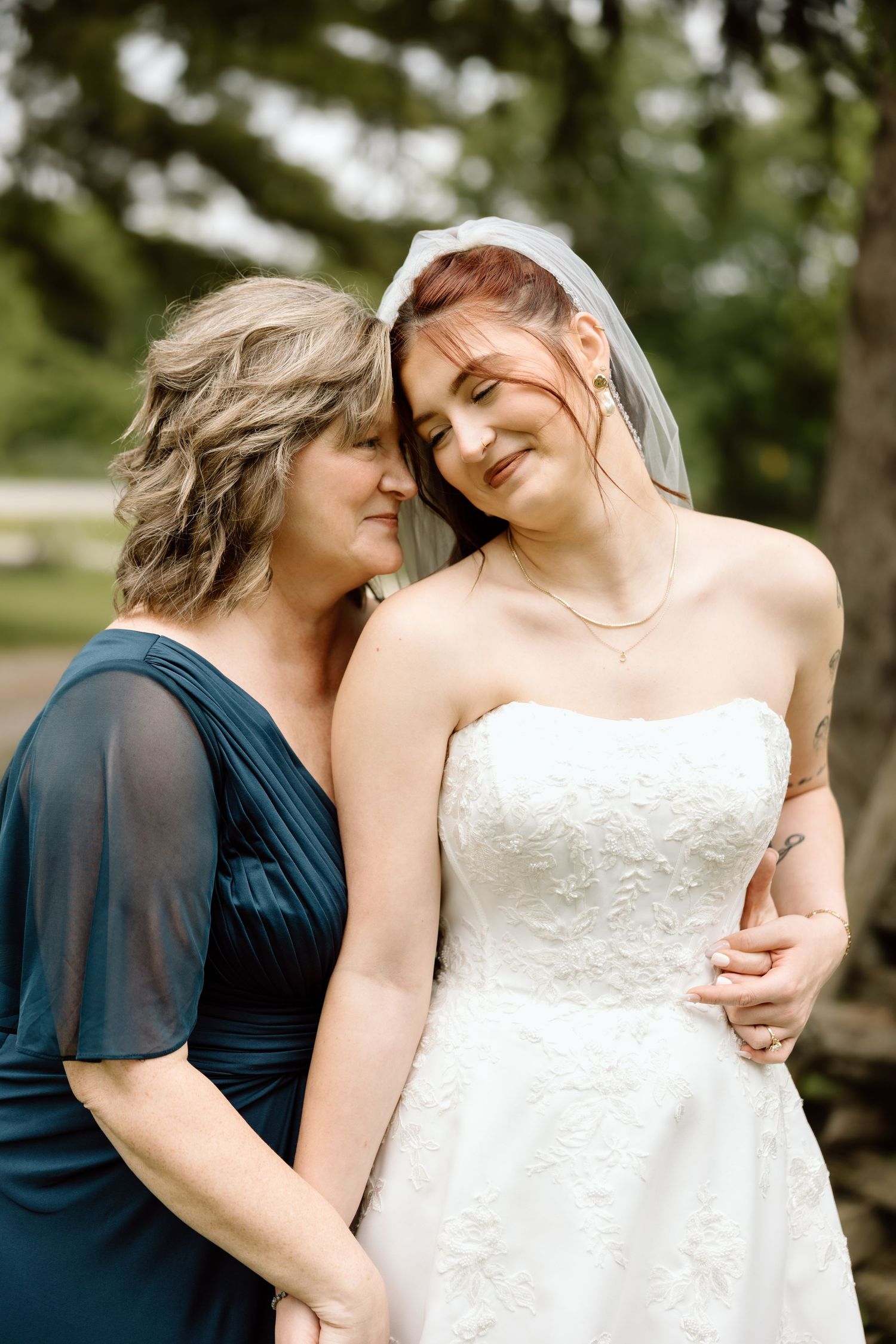 Smiling bride in lace strapless gown and veil shares tender moment with mother in teal dress.